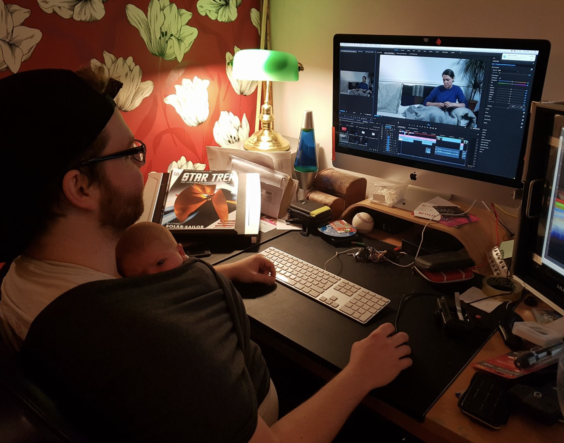 A man with glasses and a beard sitting at a cluttered desk with a baby on his lap, working on a computer editing video. The desk has various items including magazines, a keyboard, a mouse, a lava lamp, a lamp, and a baseball. The background features floral wallpaper and colorful lighting.