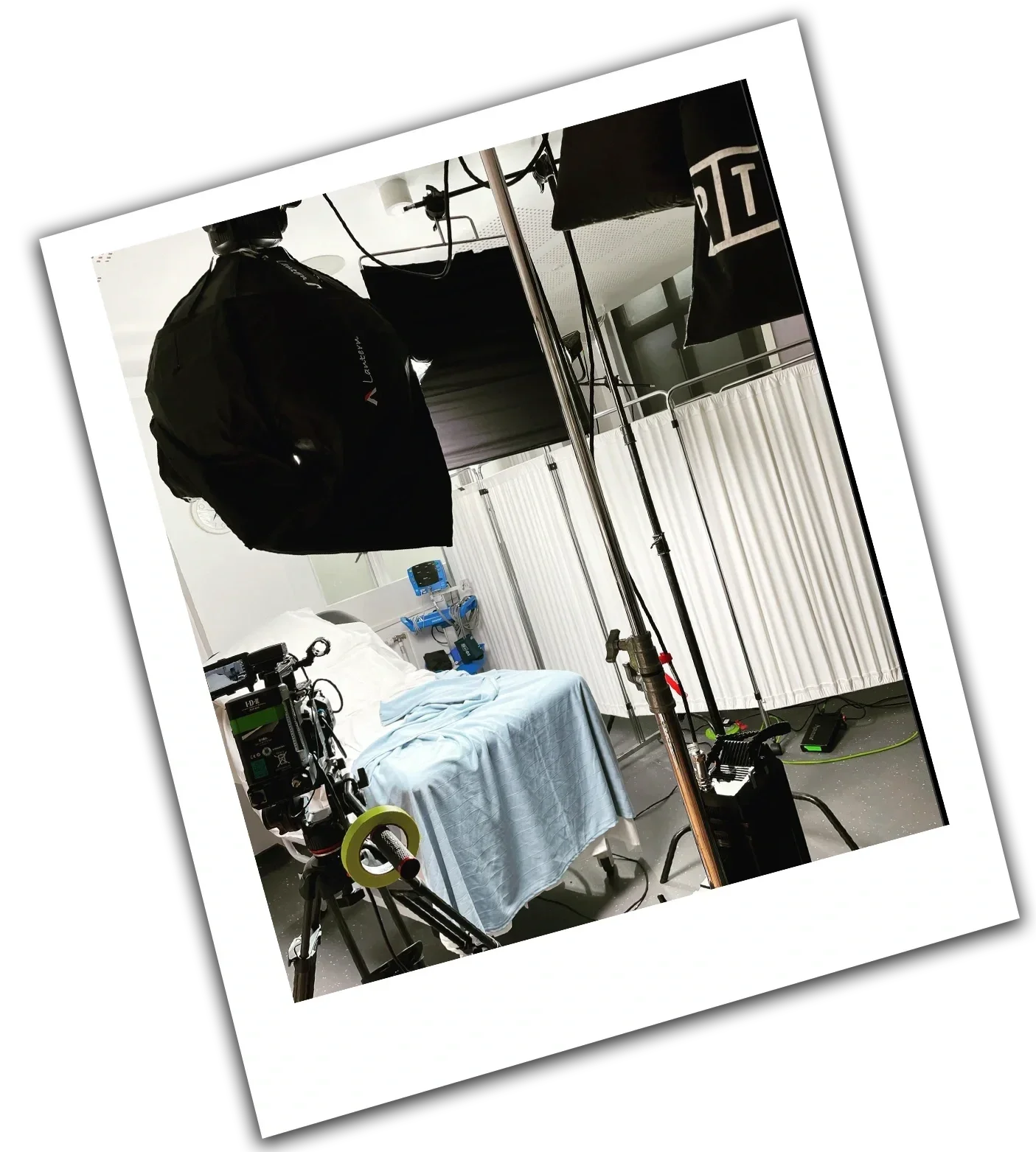 A hospital room set up for filming or photography, with a bed covered in blue sheets, medical equipment, and studio lighting equipment including softbox lights overhead.