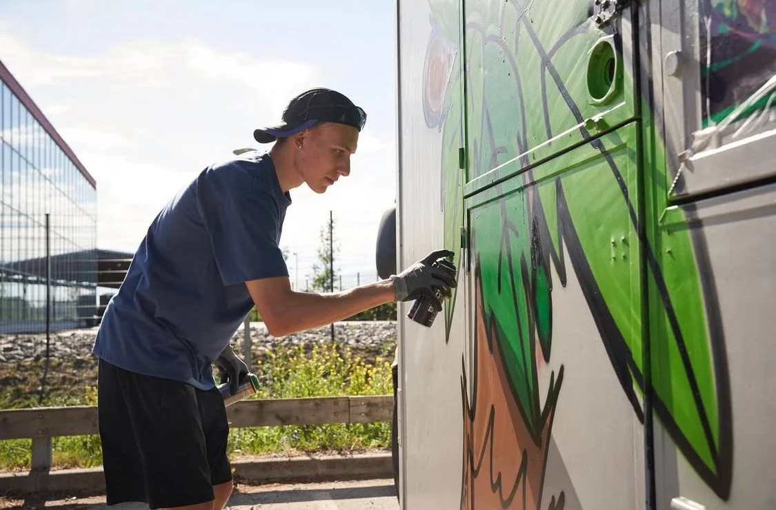 A young man spray painting graffiti on a large outdoor metal surface.