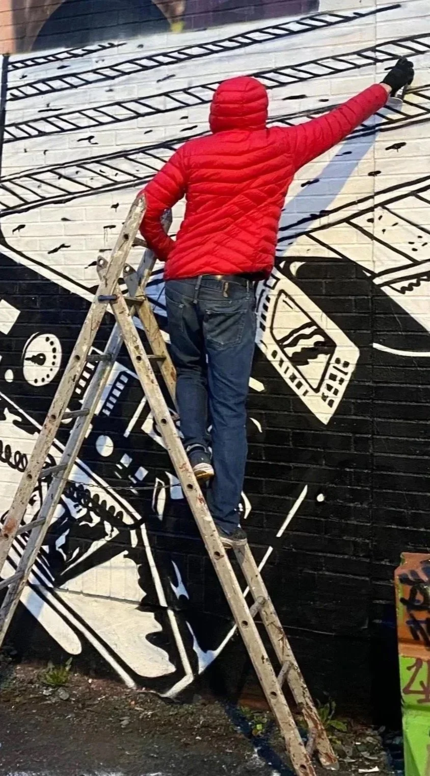 A person in a red jacket and blue jeans standing on a ladder, painting a black and white mural on a brick wall. The mural features abstract, graphic elements.