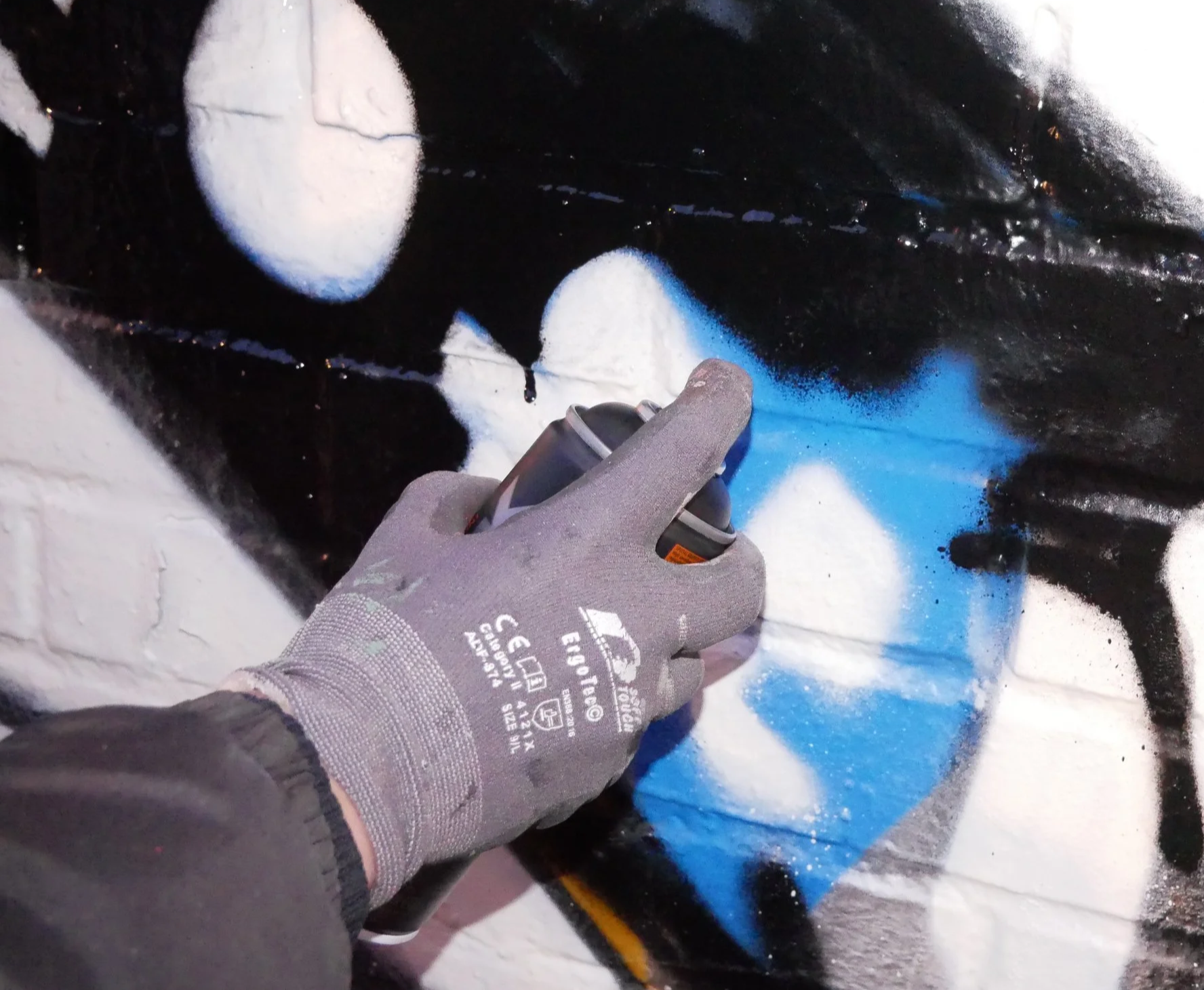 A person wearing gray gloves is spray painting a mural on a brick wall with black, white, and blue colors.
