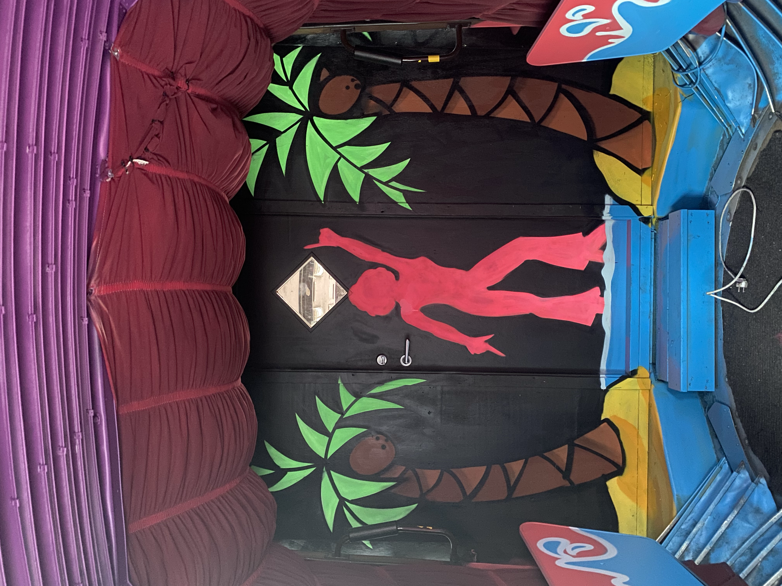 Small amusement park ride with red cushioned seats and black background wall decorated with a painted pink hiker figure, green leaves, and stylized brown trees. The ride has a blue base, and part of a blue and pink sign with the words 'Luvs' and 'Lov