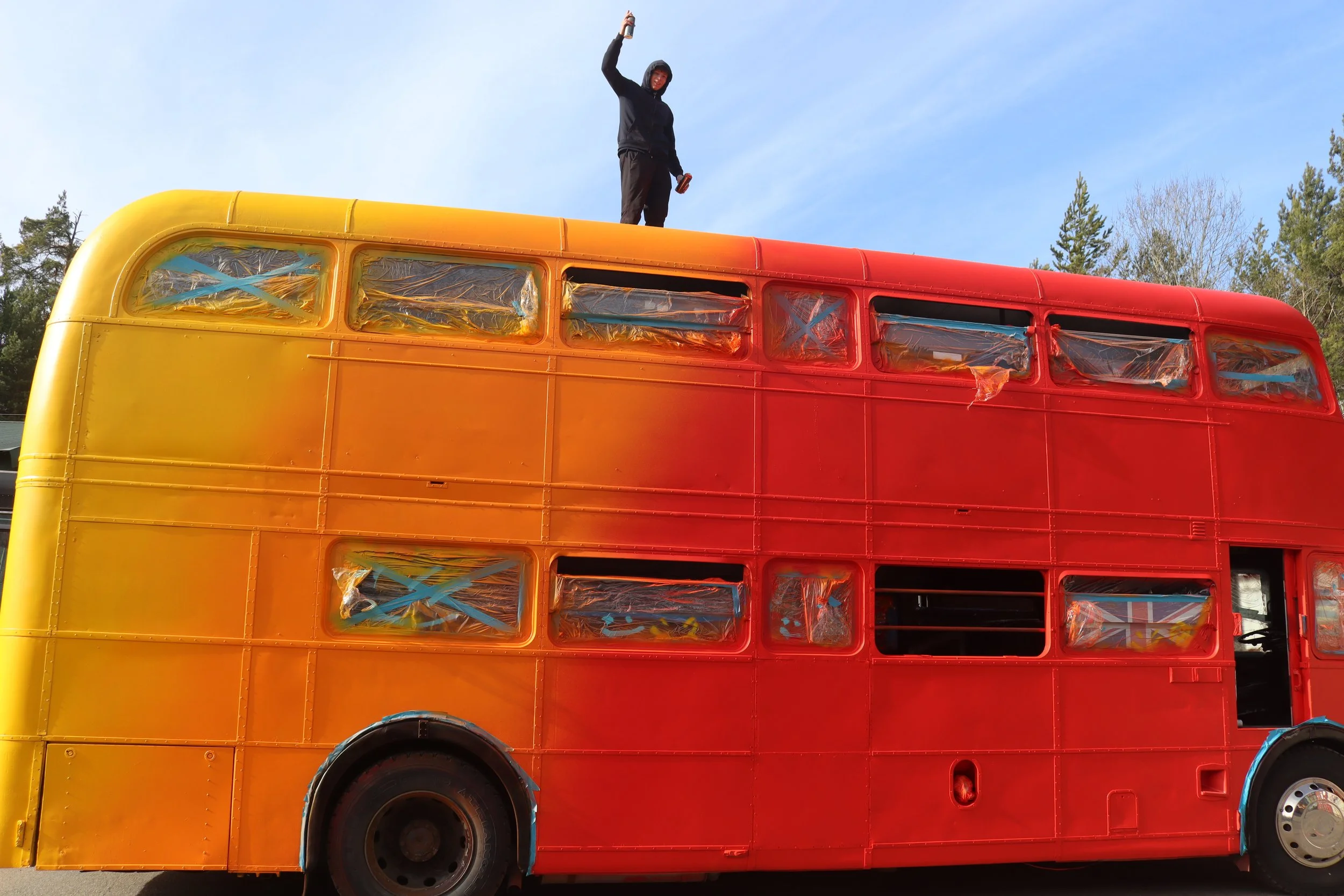 A person standing on top of a bus that is painted in a gradient of yellow to red, holding a drink, with trees and a clear blue sky in the background.