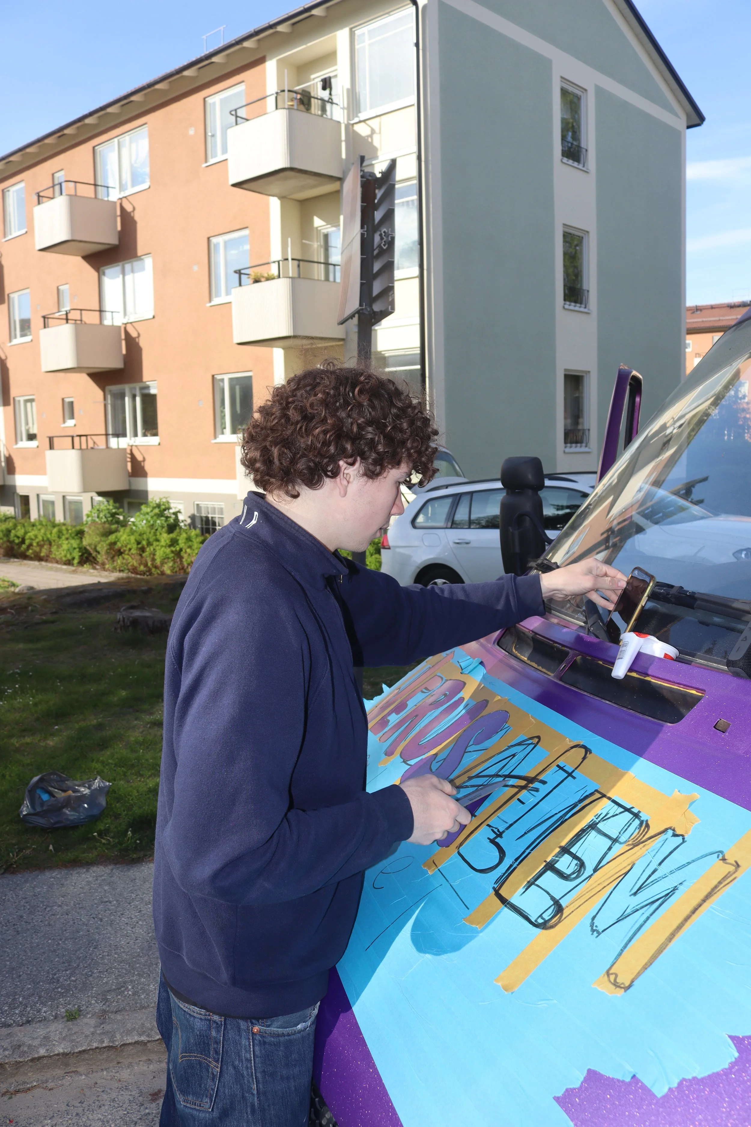 Young man creating a colorful sign on the hood of a purple vehicle, outdoors in front of residential buildings.