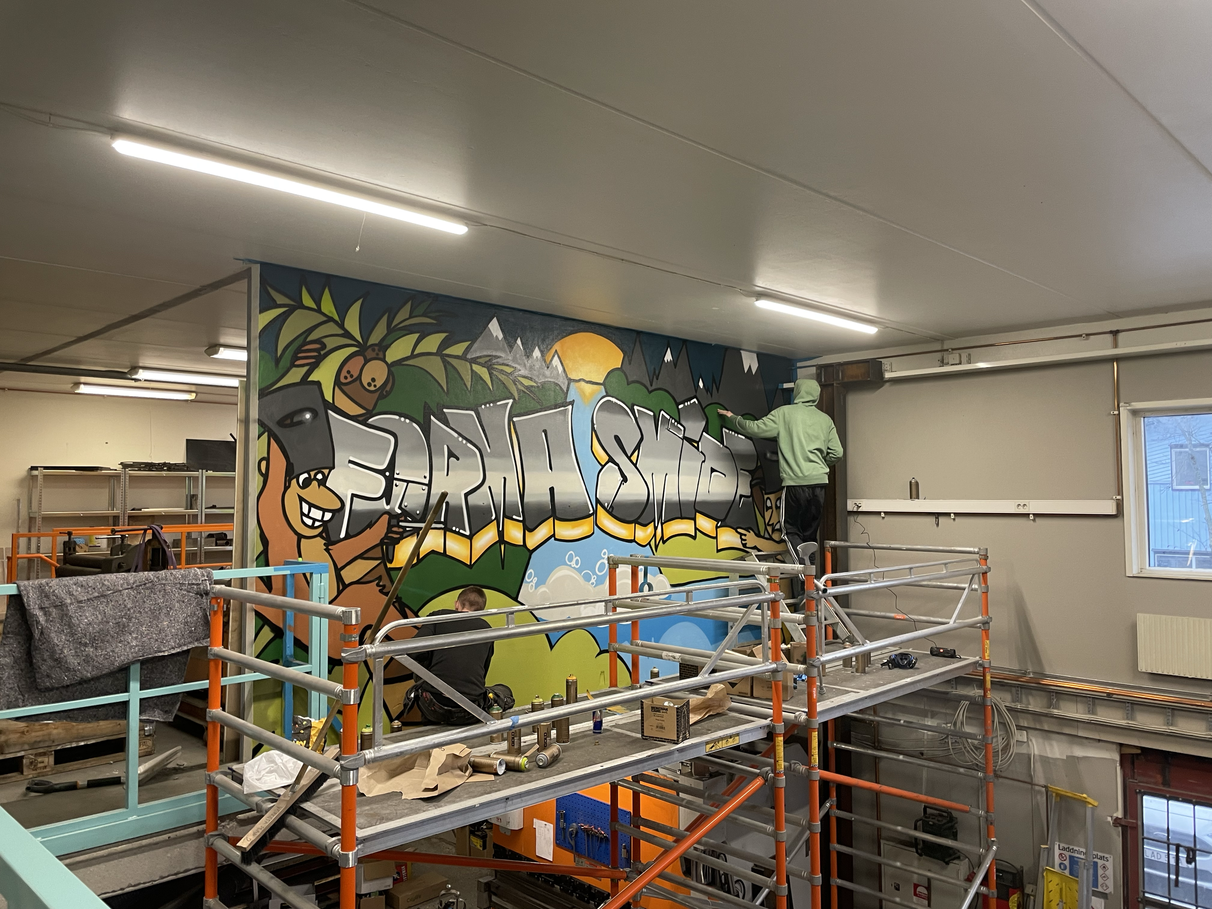 Two people painting a colorful graffiti mural on an interior wall of a building, with scaffolding and tools around.