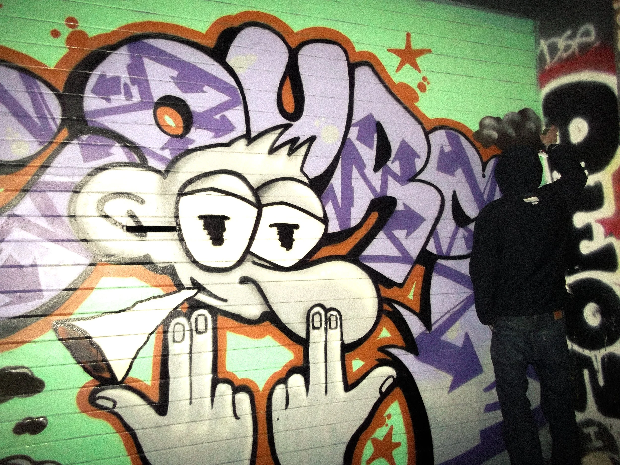 A person in a black hoodie and jeans spray paints graffiti art on a wall, which features a cartoon-style character with glasses and a finger raised.