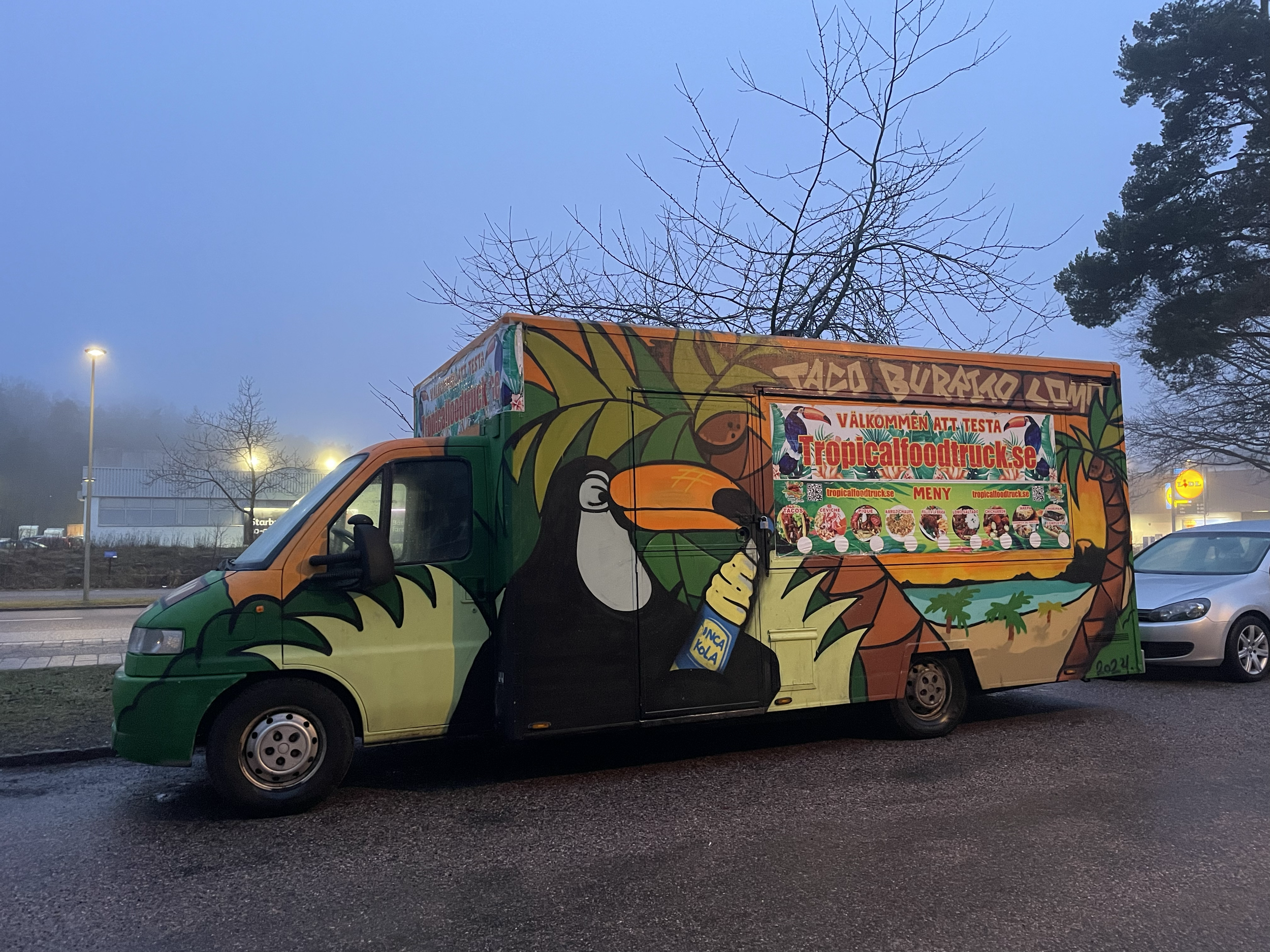 Colorful food truck with tropical design, featuring toucans, leaves, and a cocktail glass, advertising a taco and burrito menu from tropicalfoodtruck.se.