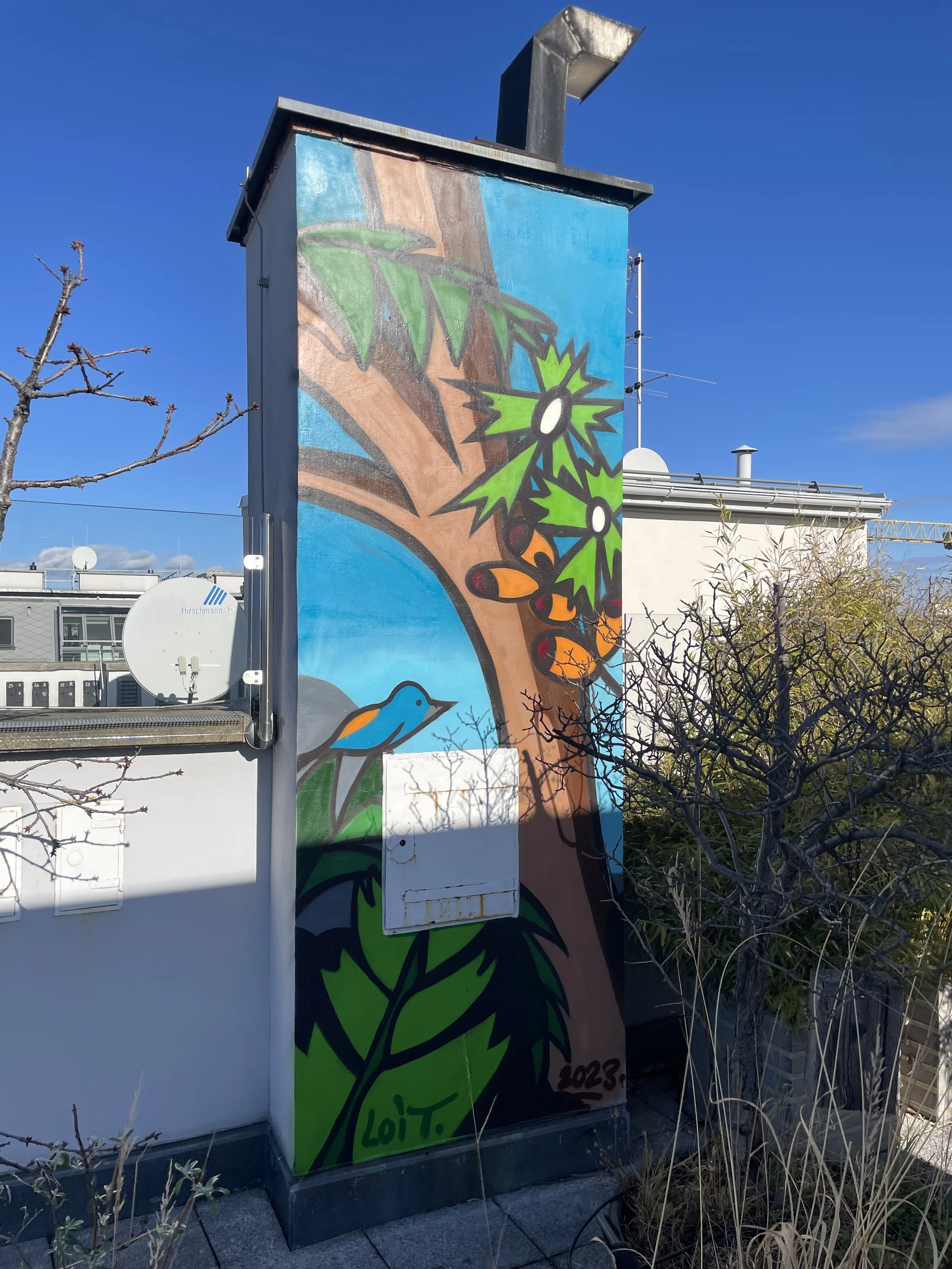 A colorful mural on a utility box depicts a tree with green leaves, orange and black insects on the branch, and a bluebird with orange wings on the lower left. The background is a bright blue sky, and the mural is signed with the artist's initials an