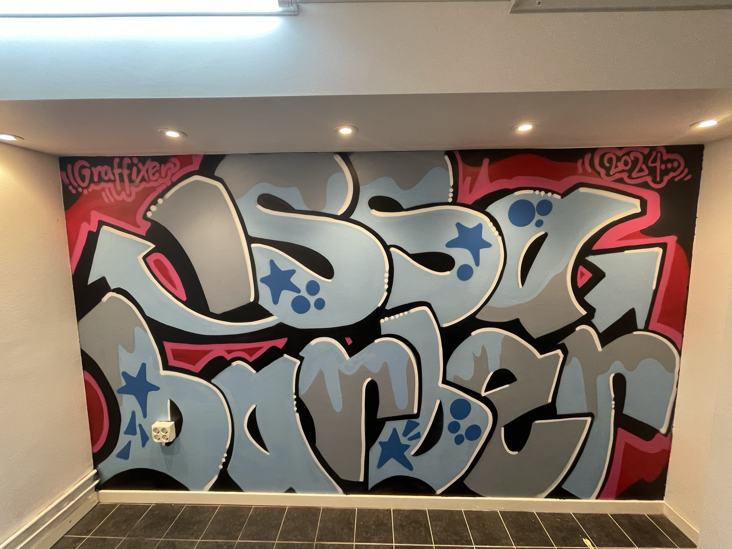 Colorful graffiti mural with abstract shapes and stars, signed with 'Graffiti' in the top left corner and '2024' in the top right corner, on an interior wall.