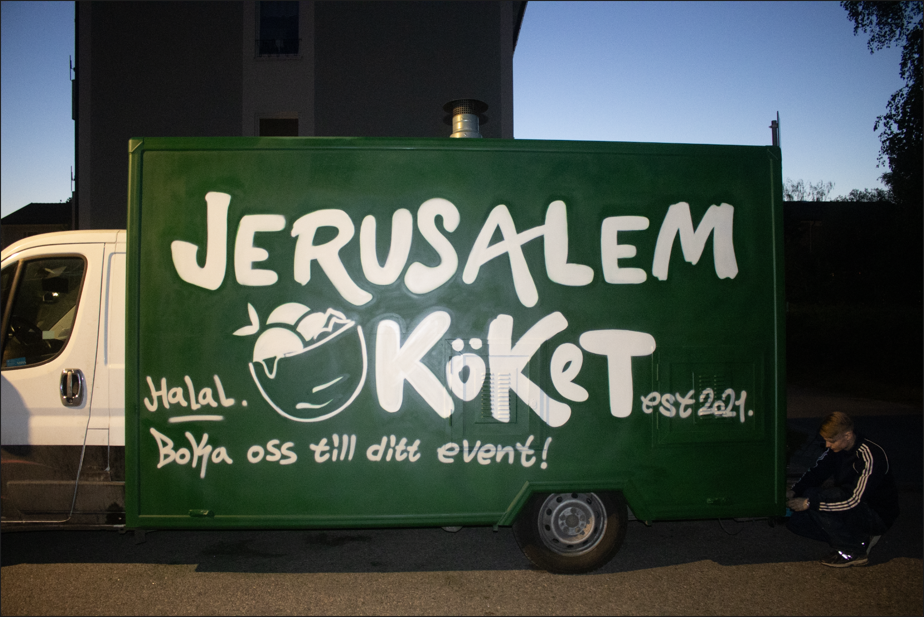 Green food truck with white handwritten text advertising Jerusalem Kiosk, a halal food stand established in 2021, with a slogan in Swedish inviting customers to join their event. A man kneels beside the truck adjusting something near the wheel, and a