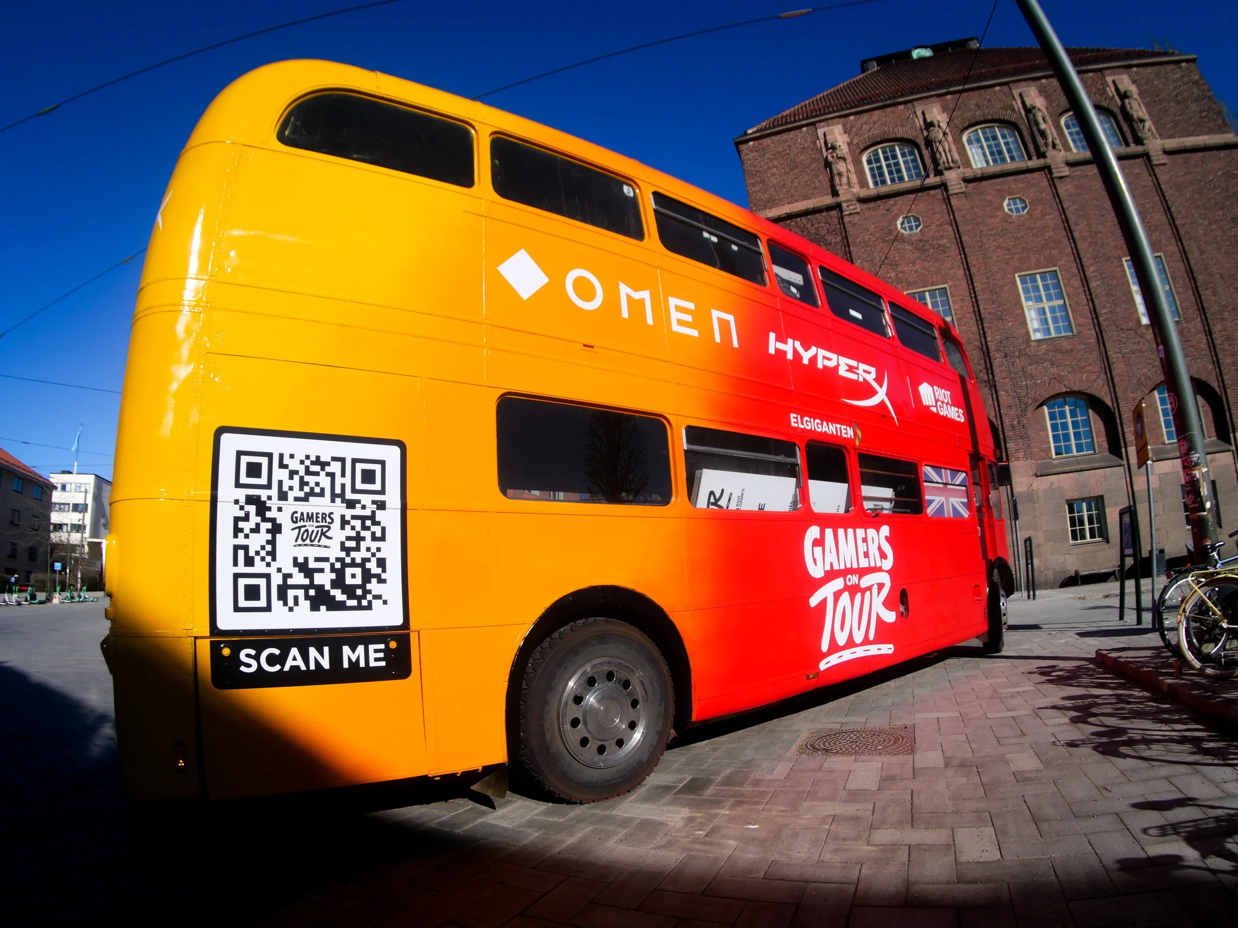 A colorful double-decker bus painted in red and yellow, featuring logos of OMEN and HyperX, with a QR code and the text 'Scan Me' on the side, parked on a city street with historic brick buildings and bicycles nearby.