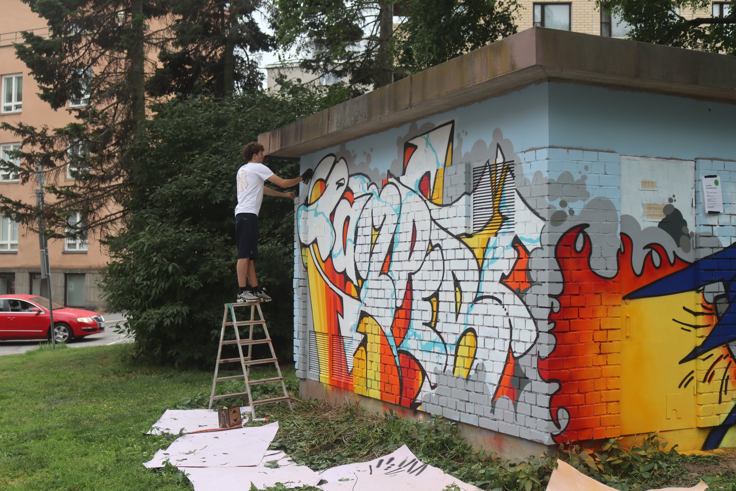 A young man standing on a ladder painting a colorful graffiti mural on the side of a small brick building in an urban park.