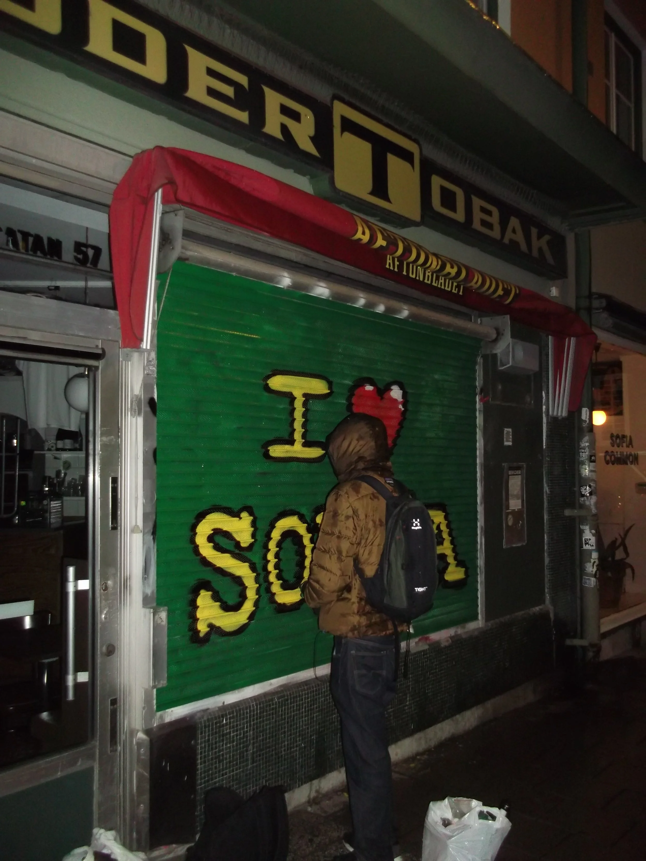 A person with a backpack standing outside a closed storefront with a green shutter painted with the words 'I ♥ SO'. The store has a sign above that appears to read 'DERT OBAK' and a red canopy, possibly at night.