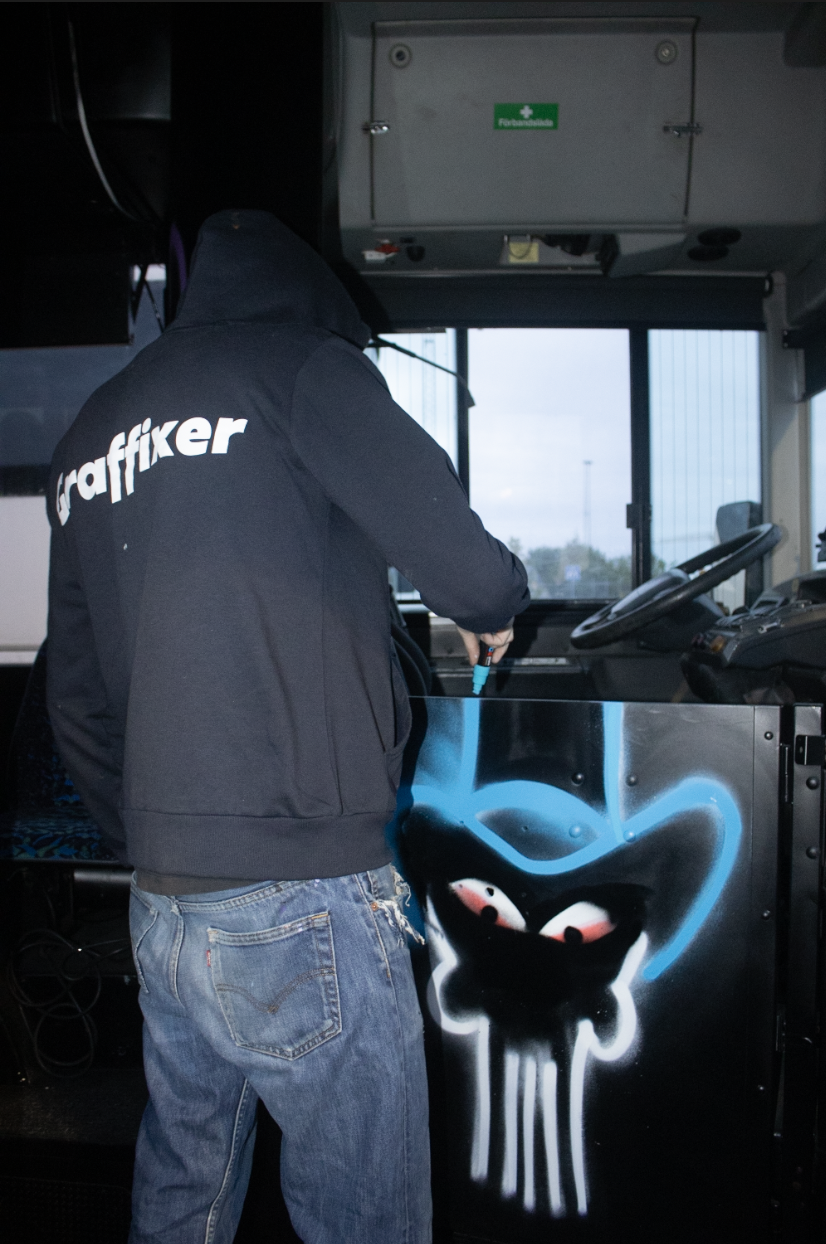 A graffiti artist wearing a black hooded jacket with 'graffixer' written on the back, spray painting a cartoonish face on a black surface inside a vehicle. The face features large eyes and a wide-open mouth with drips of paint beneath it.