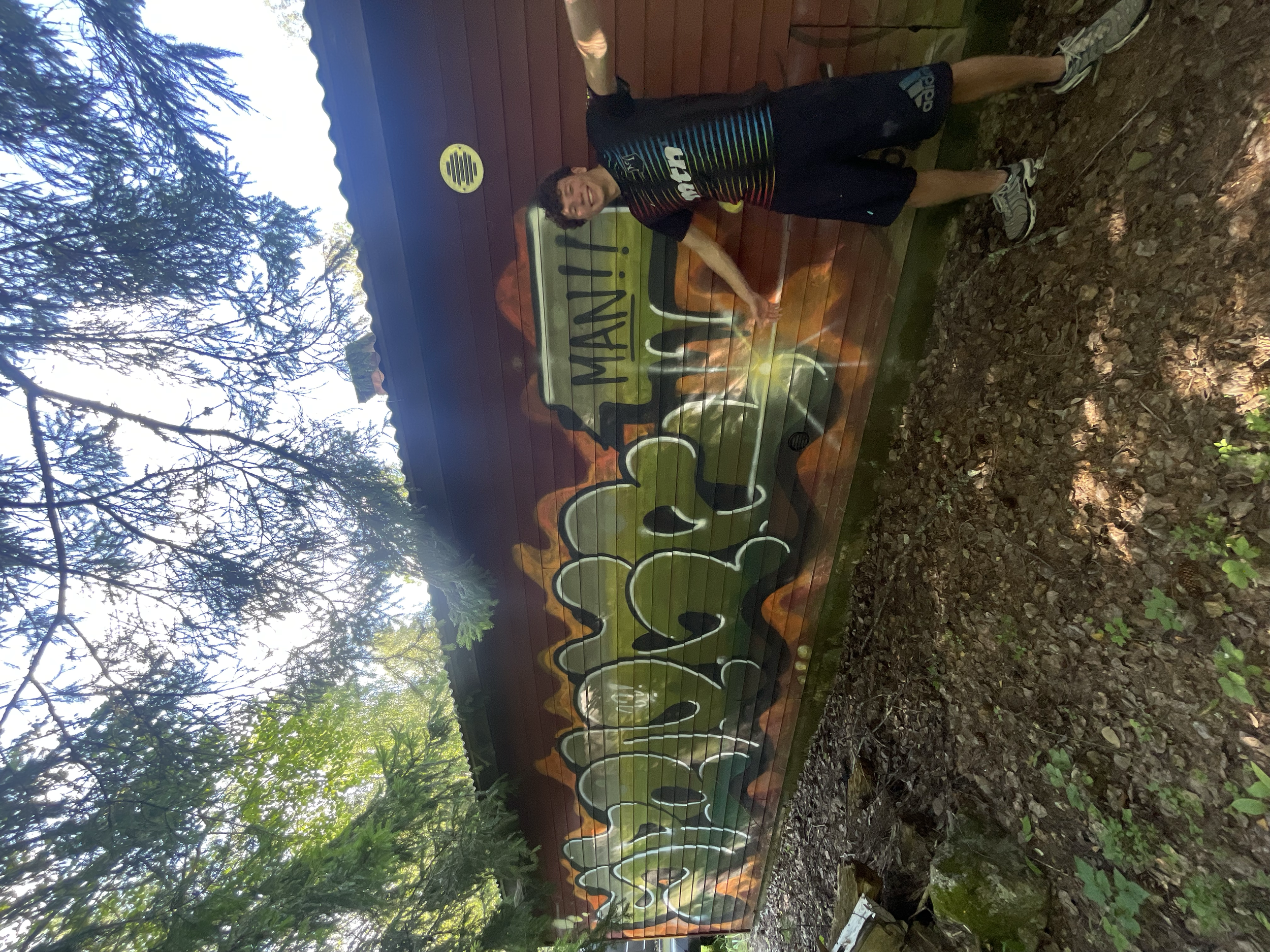 A person in athletic clothing is standing next to a colorful outdoor sign or mural with the words "Fighting Manji" and a dragon design, outdoors surrounded by trees and dirt ground.