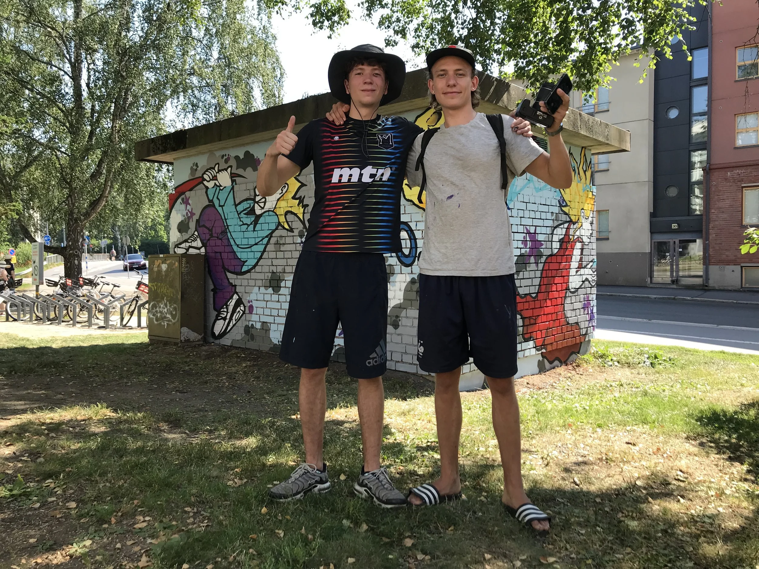 Two boys standing outdoors in front of a colorful graffiti wall, smiling and giving thumbs up. One boy is wearing a black hat, striped shirt, and black shorts, while the other is wearing a gray shirt, black shorts, and flip-flops.