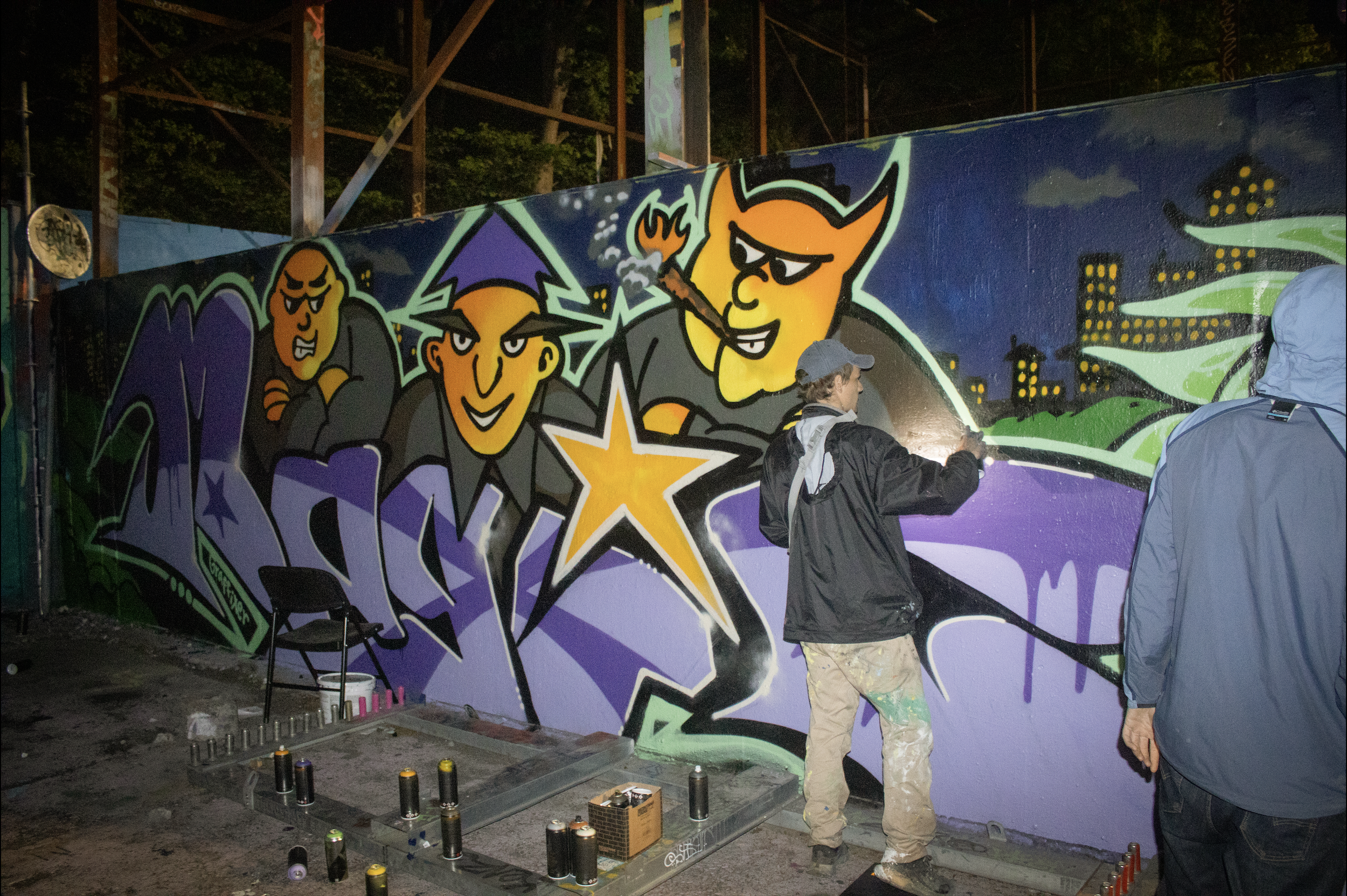Two artists create colorful graffiti on a large outdoor wall at night. One artist sprays paint, and the other stands nearby. Spray paint cans and supplies are on the ground.