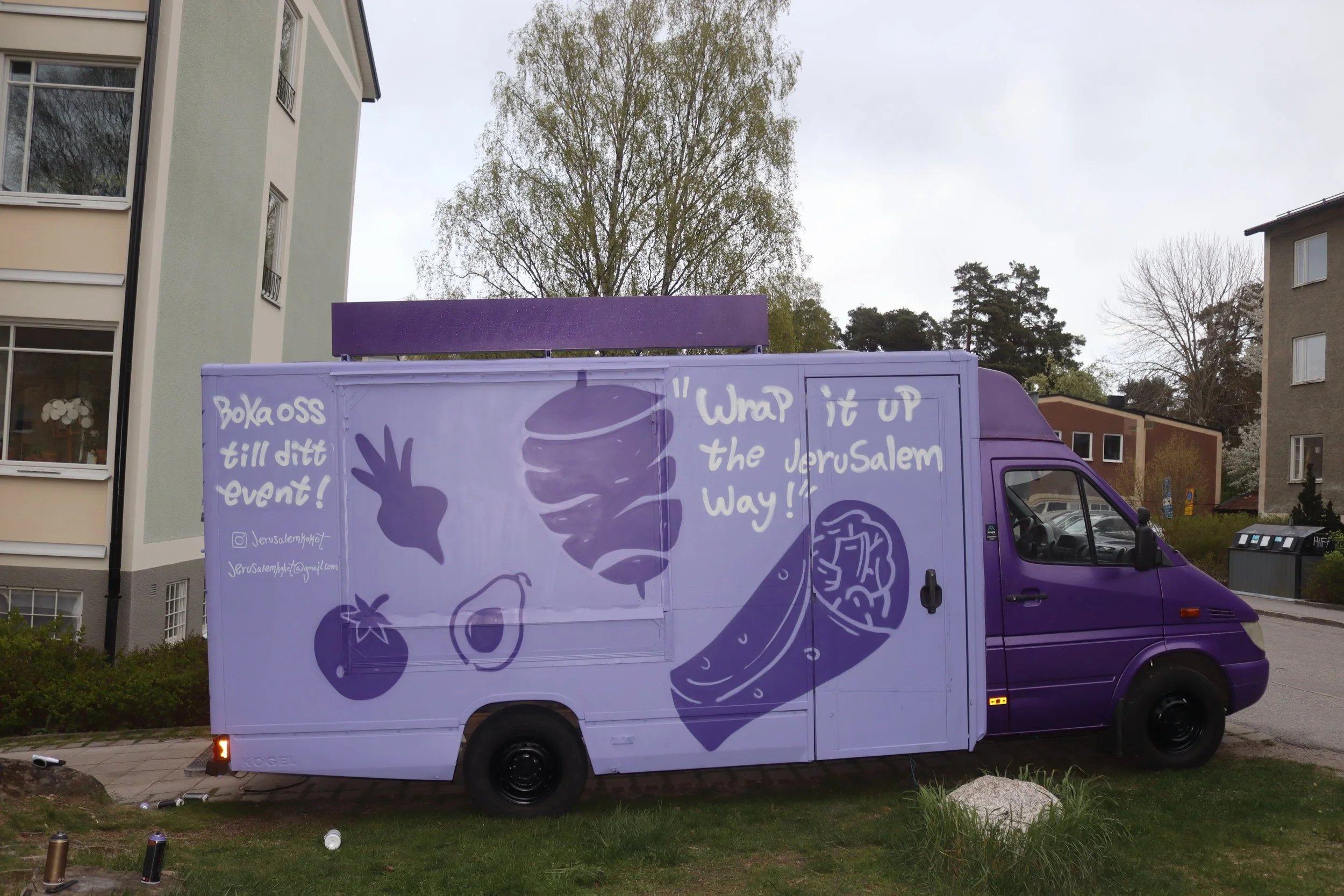 A purple food truck parked on a grassy area with apartment buildings and trees in the background. The truck features handwritten-style artwork and text supporting Jerusalem, with illustrations of a hand, tomato, onion, and a wrap with a brain inside.