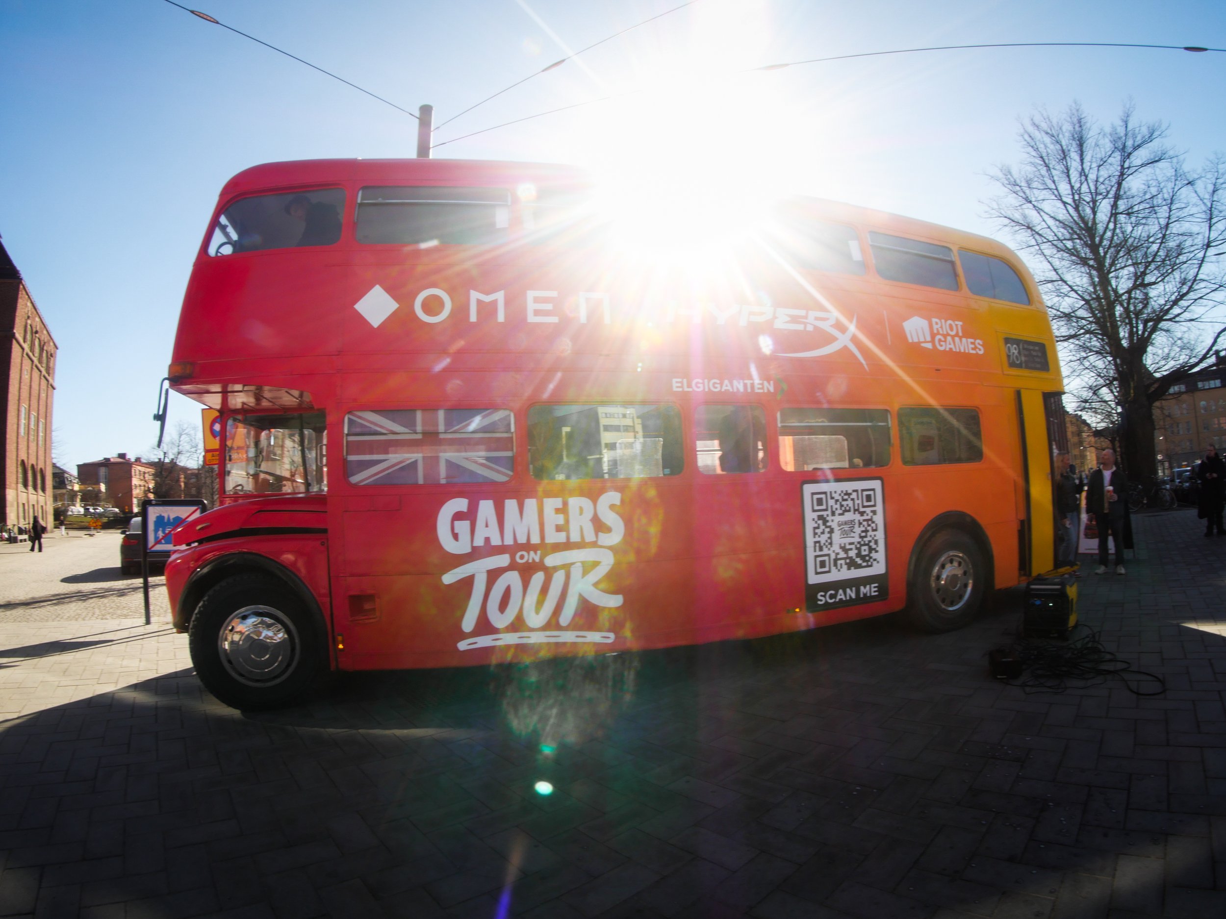 A red double-decker bus with gaming advertisements and a QR code parked on a city street on a sunny day, with a tree and buildings in the background.