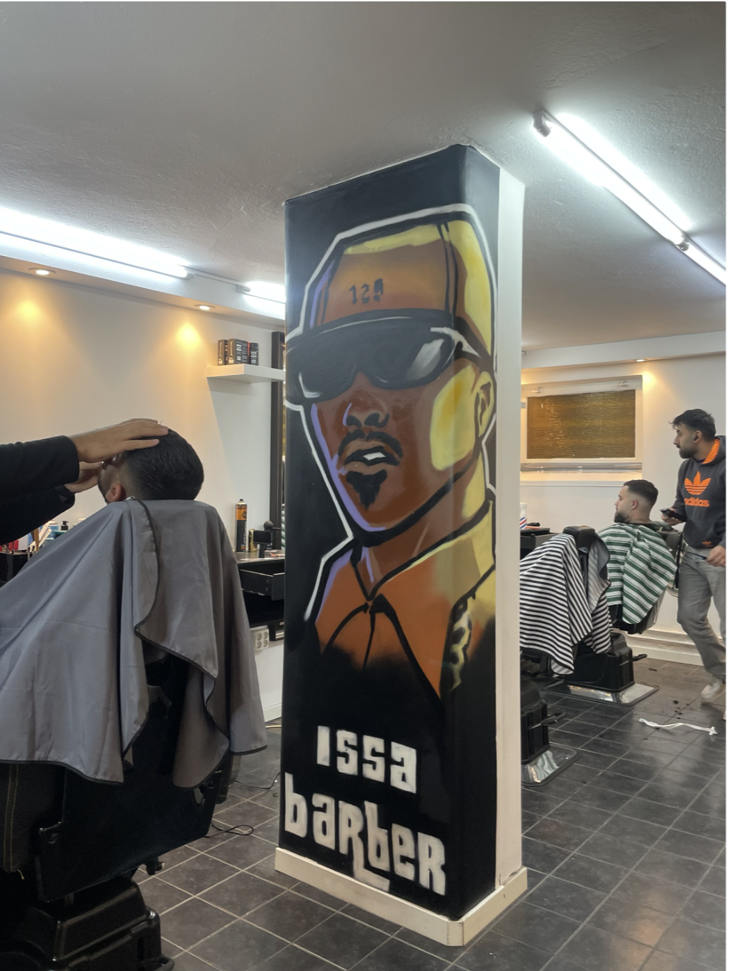 A barber shop with a colorful mural of a man wearing sunglasses and a cap labeled '1553 Border' on a column. A customer is getting a haircut, and another person in a black Adidas hoodie is standing nearby.