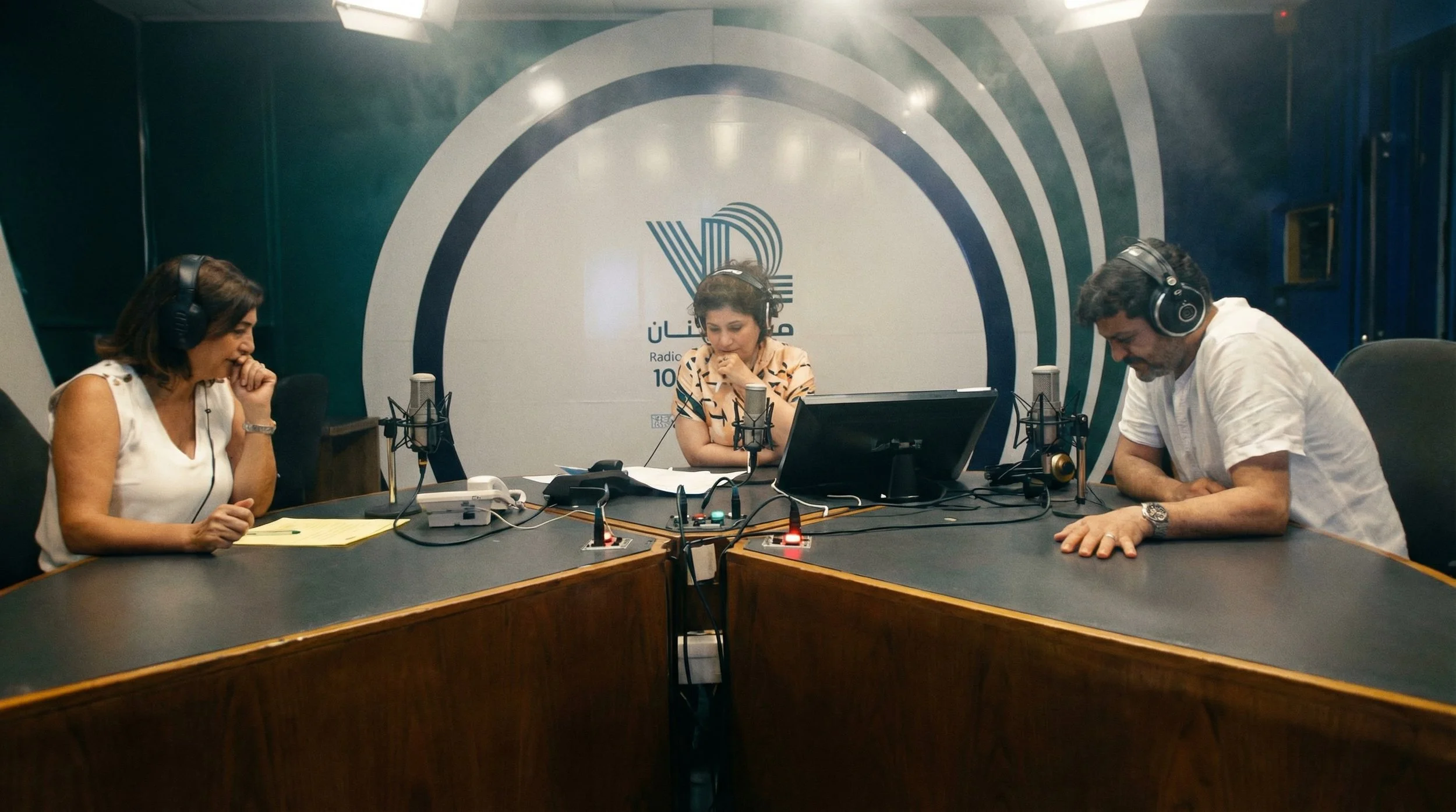 Three people wearing headphones are sitting at a table in a radio studio, with microphones, a computer monitor, and recording equipment, in front of a wall with a logo and Arabic text.