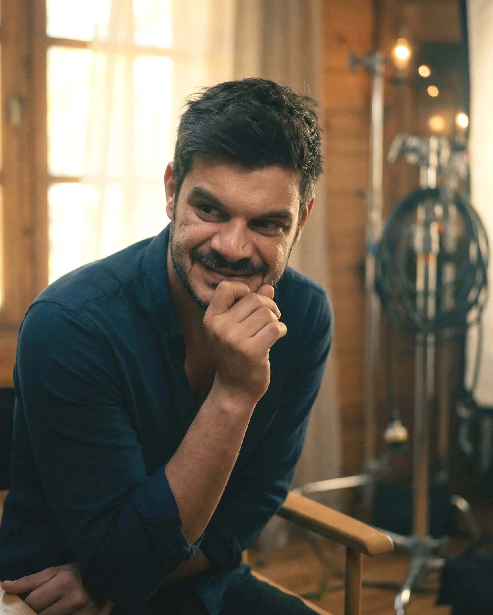 A man with dark hair and a beard, wearing a dark blue shirt, sitting in a wooden room with natural light coming through the window, resting his chin on his hand and smiling. Julien Maalouf