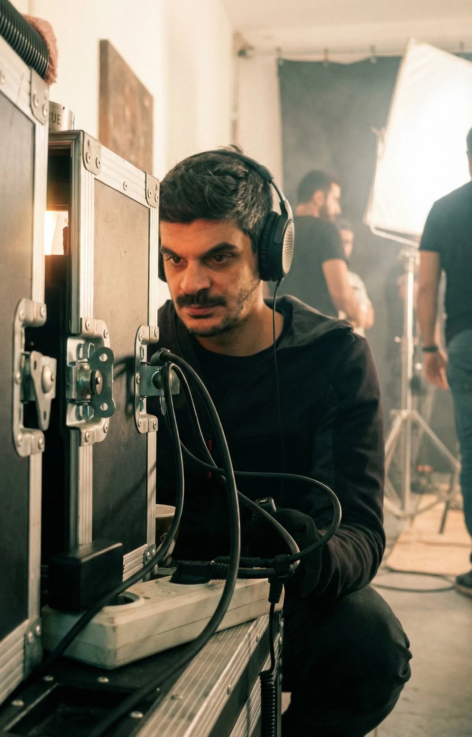 A man with headphones working on electronic equipment on a film or photography set with crew members and studio lighting in the background. Julien Maalouf