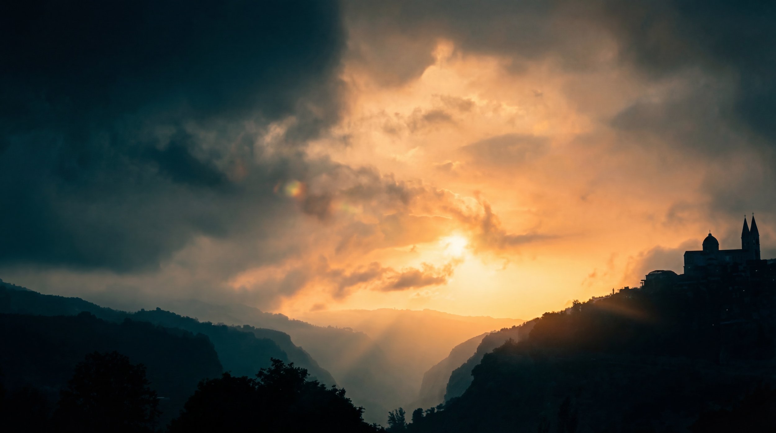 Sunset over a valley with dark hills and a silhouette of a church with two spires on a hill on the right. Julien Maalouf