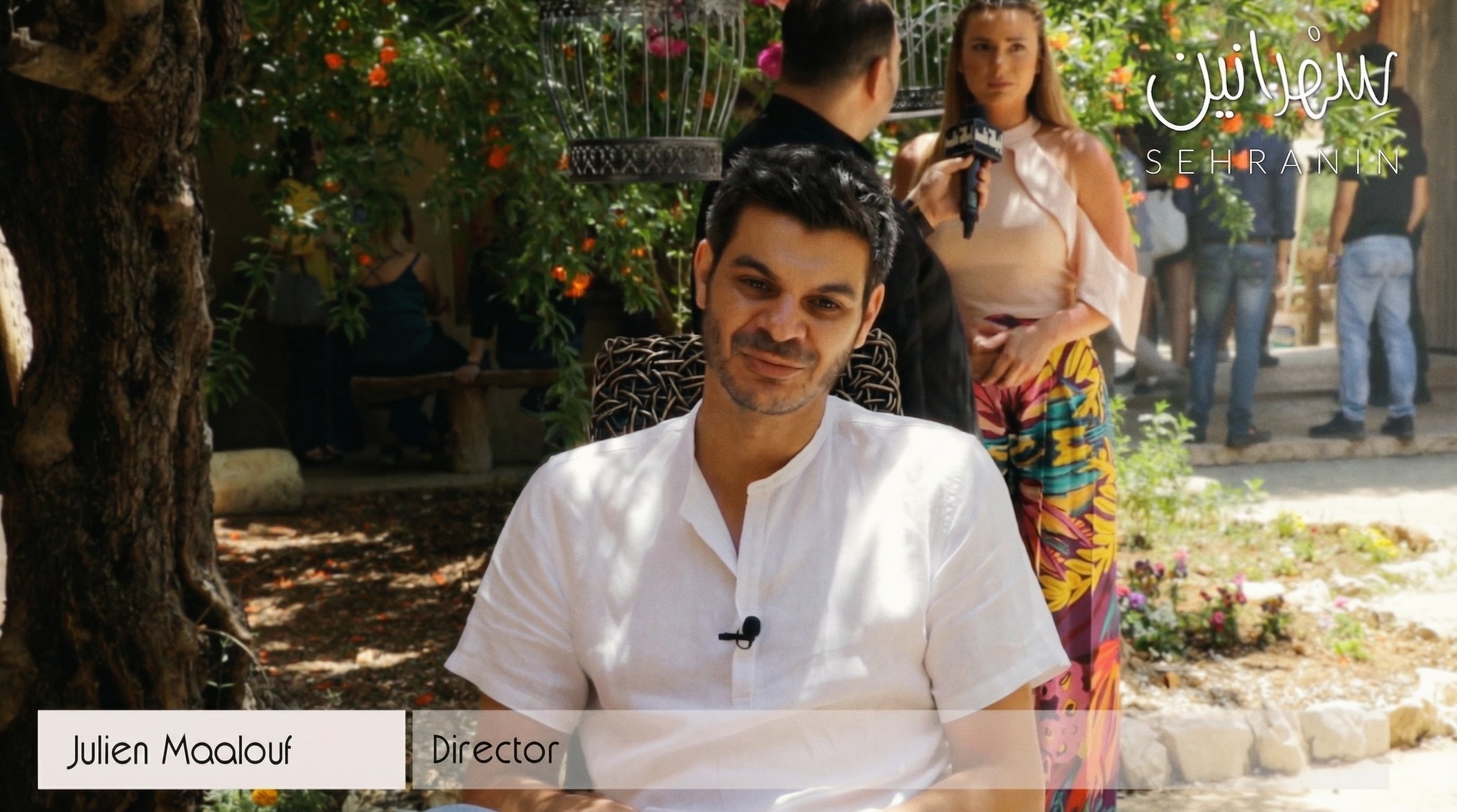 A man sitting outdoors with a white shirt and a microphone clipped to his shirt, smiling at the camera, with a woman and a man in the background, and several other people standing nearby. There are colorful flowers and trees around.