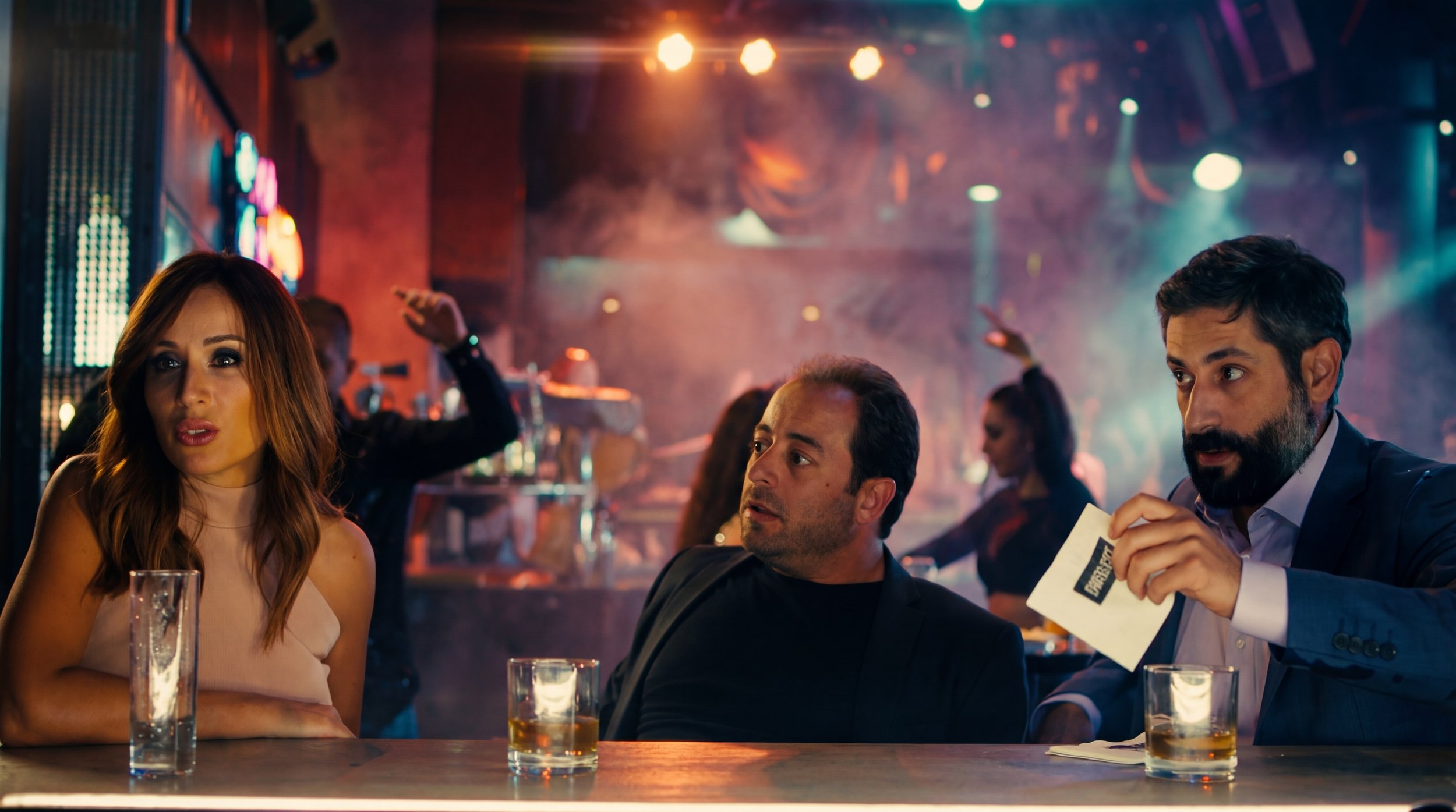 Three people sitting at a bar, talking and drinking. The setting is a lively nightclub with colorful lights and other patrons in the background.