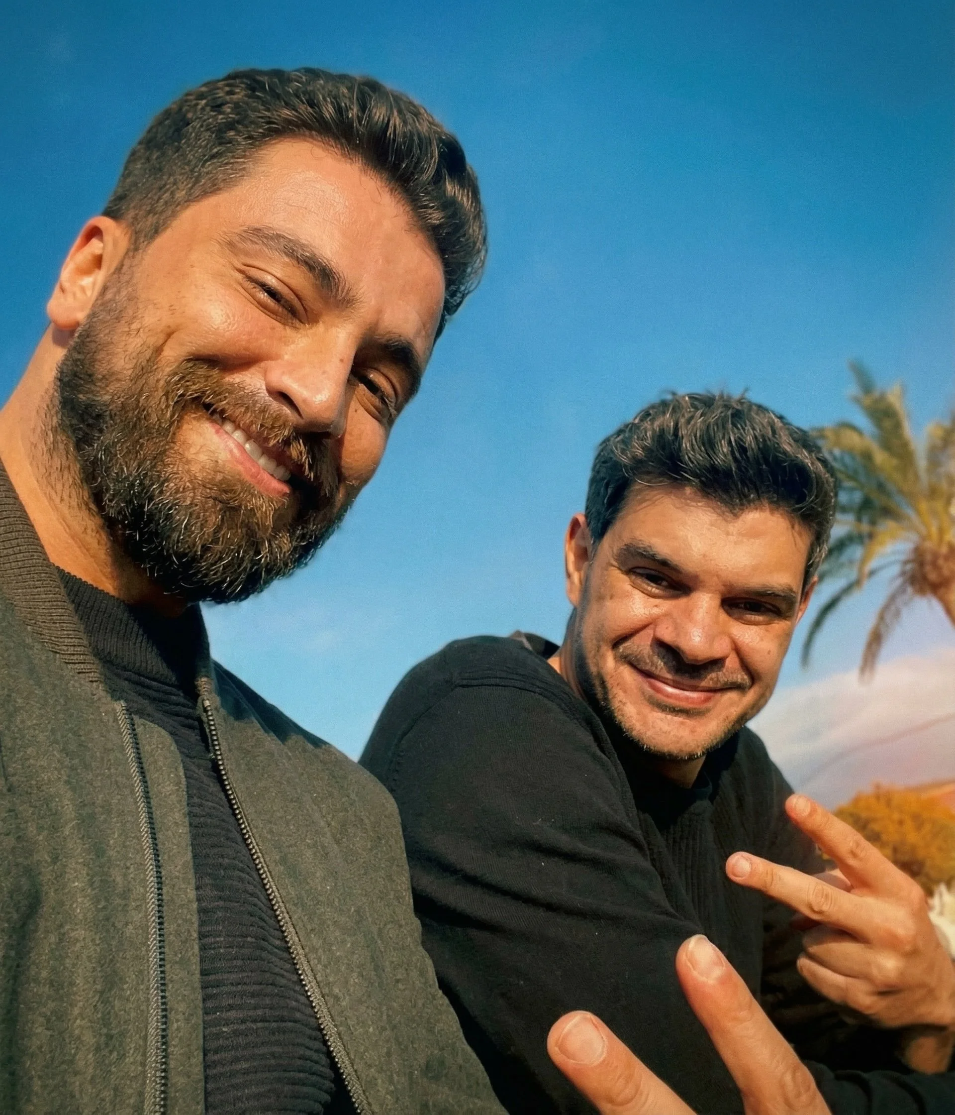 Two smiling men taking a selfie outdoors with clear blue sky and palm trees in the background. One man has dark hair and a beard, wearing a dark jacket. The other man has dark hair, no beard, and is making a peace sign with his fingers. Julien Maalou