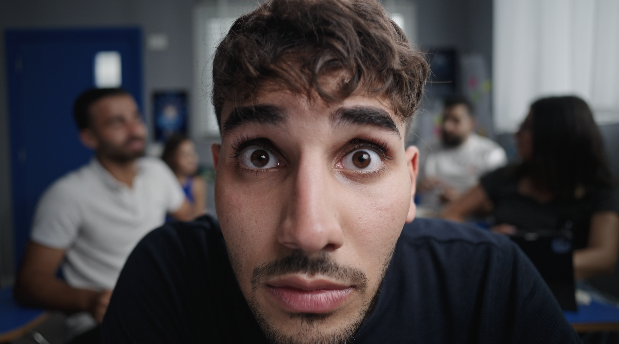 Close-up of a young man with brown hair and facial hair looking directly at the camera with wide eyes, in a classroom setting with blurred students in the background. Julien Maalouf