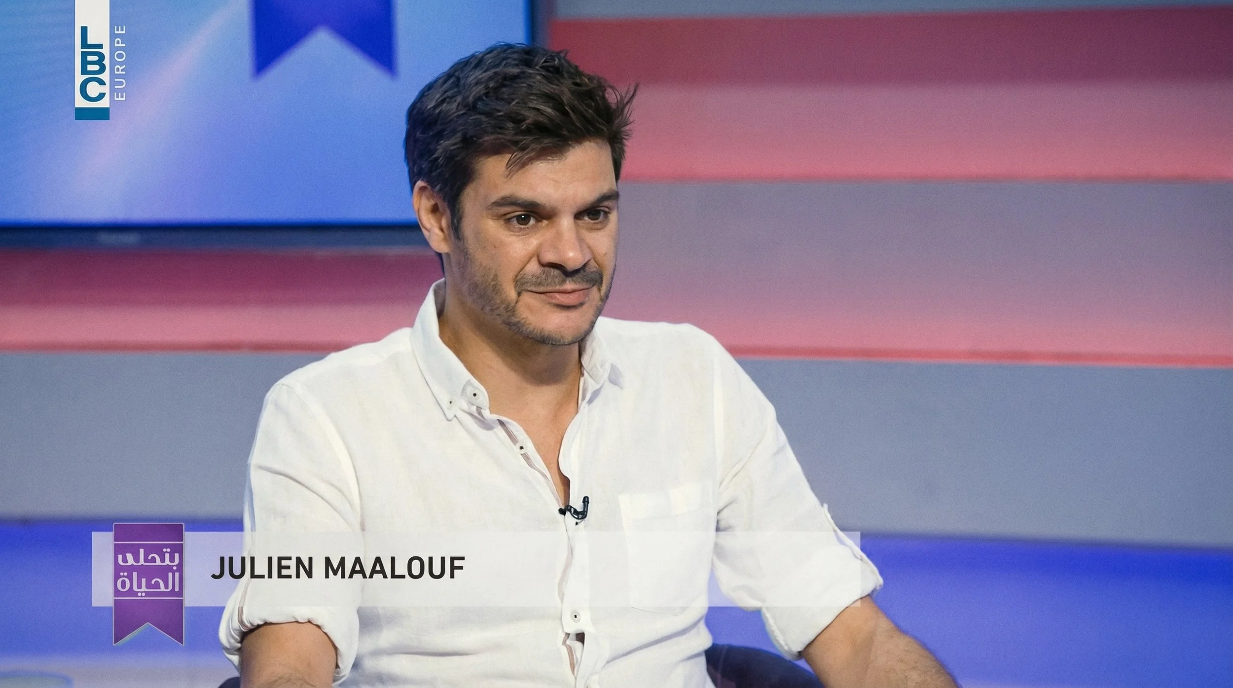 A man with dark hair and a light beard, wearing a white button-up shirt, sitting on a TV set with colorful horizontal stripes background, and a logo on the top left corner that says 'LBC EUROPE.'