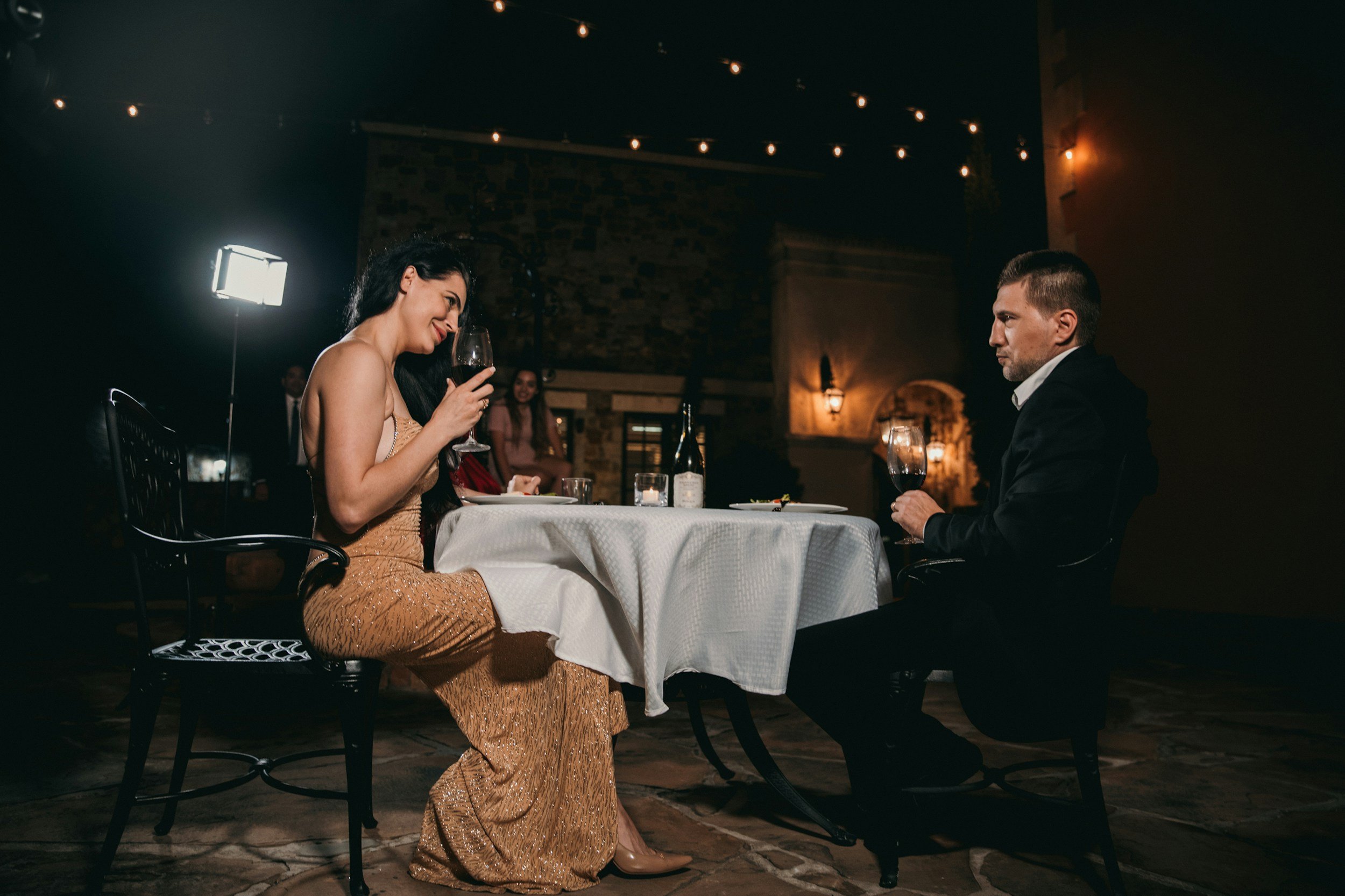 A woman in a gold dress and a man in a tuxedo sitting at a dinner table in a dimly lit restaurant, engaging in conversation with glasses of wine.