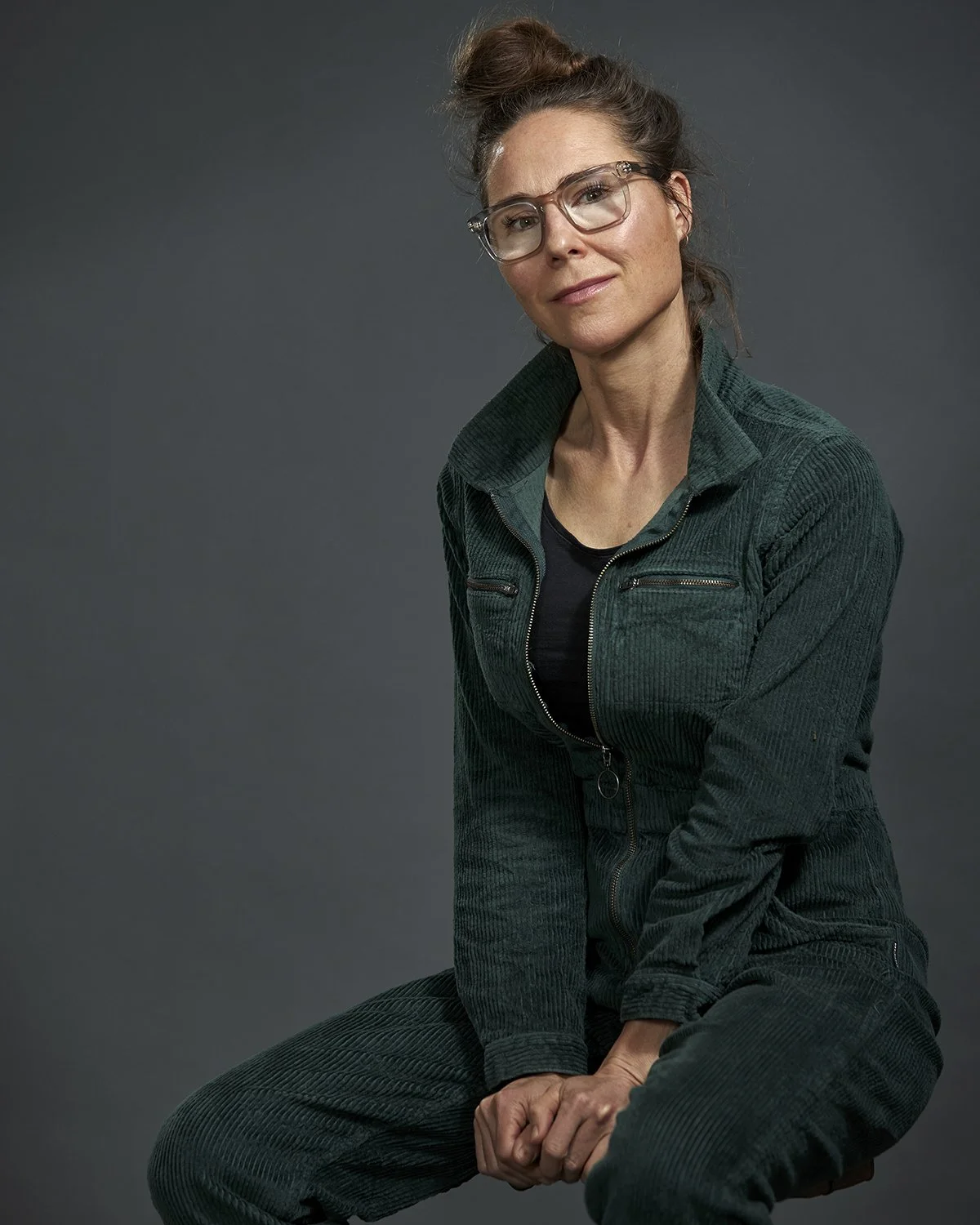 A woman with glasses and curly hair in a messy bun wearing a green corduroy jacket sitting against a plain dark background.