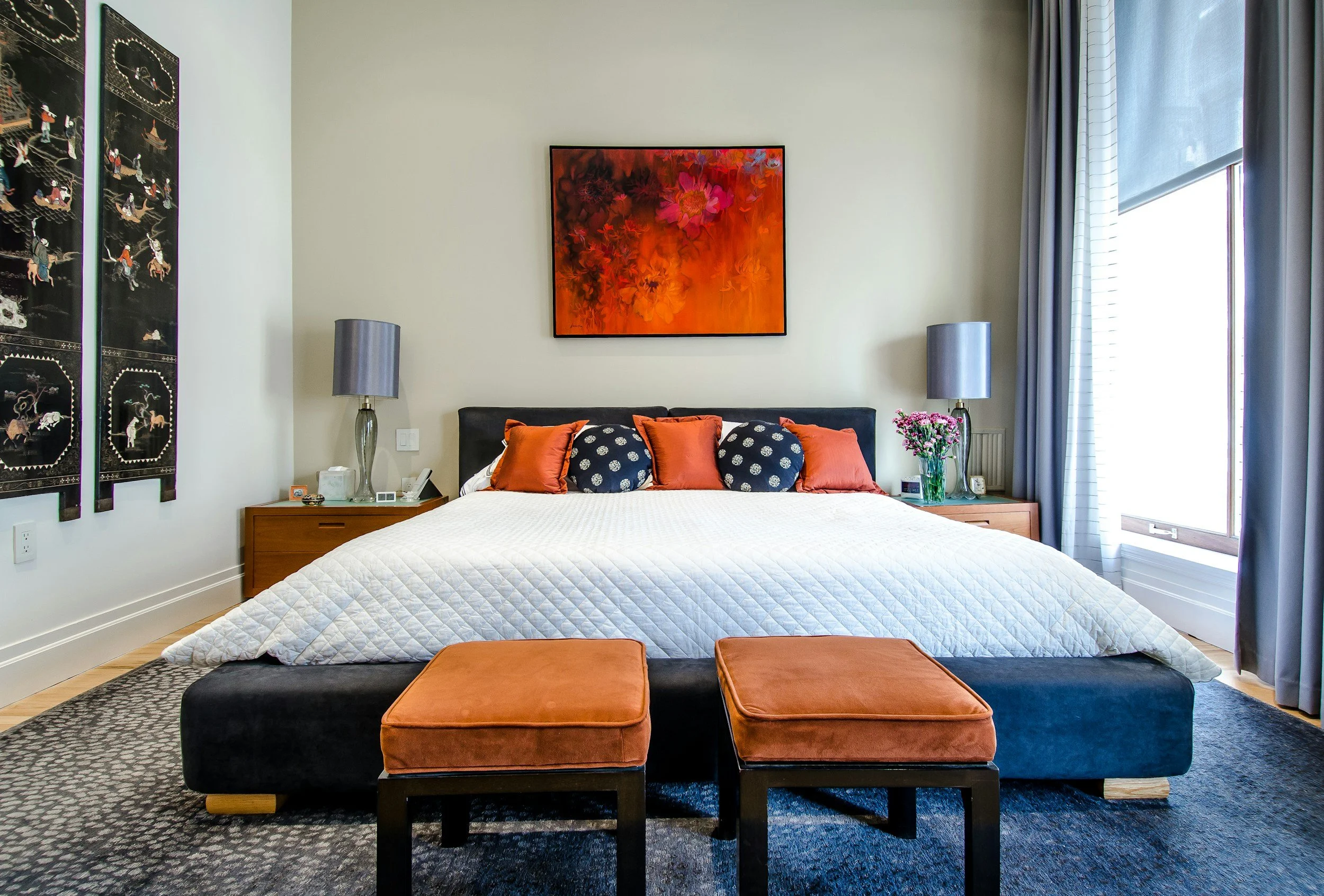 A modern bedroom with a large bed, decorative pillows, and two bedside tables with lamps. A framed abstract floral artwork hangs above the bed, and a window with blue curtains is on the right. Two orange ottomans are at the foot of the bed, and a dark rug covers the floor.