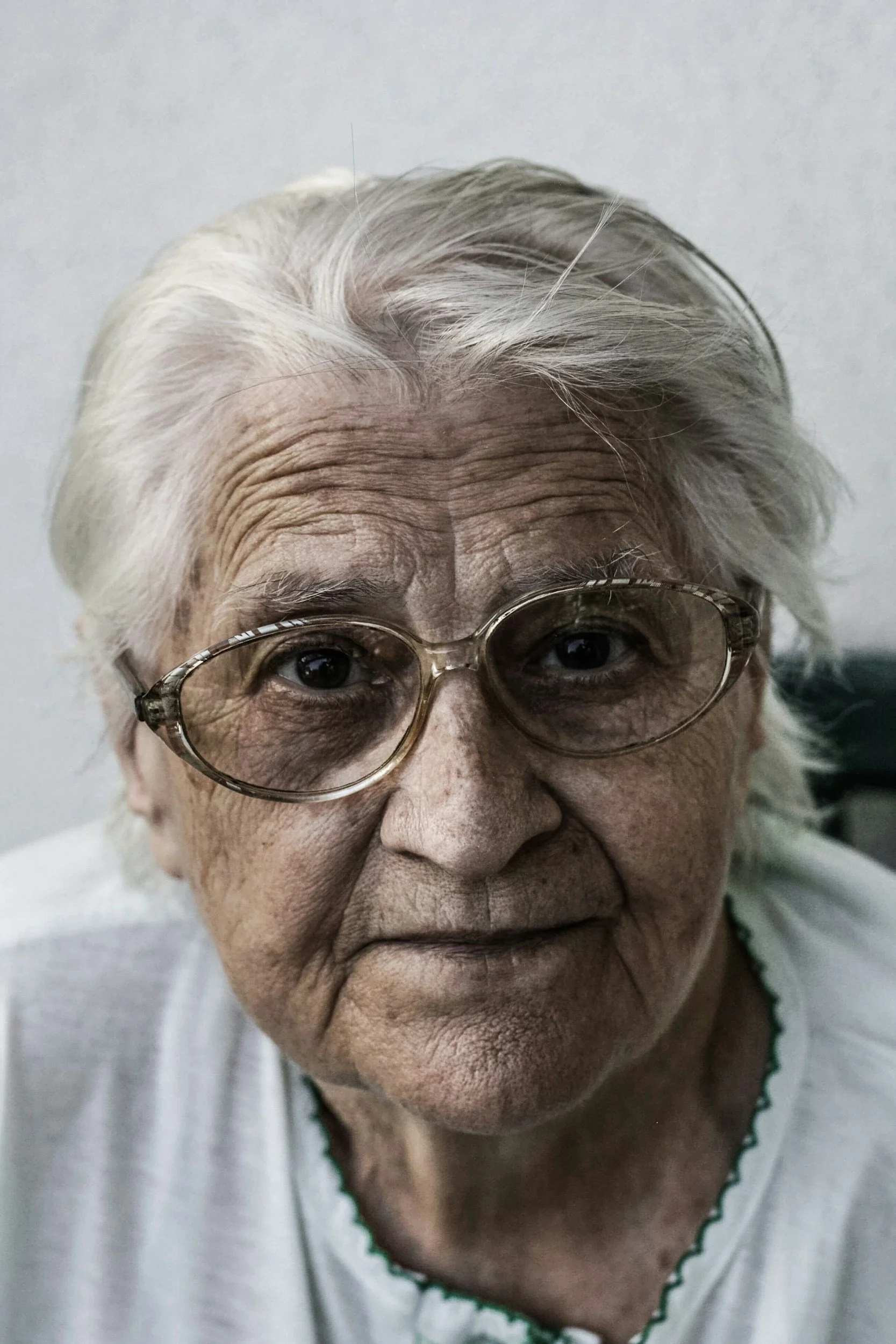 Close-up of an elderly woman with white hair and glasses, showing facial wrinkles and signs of aging.