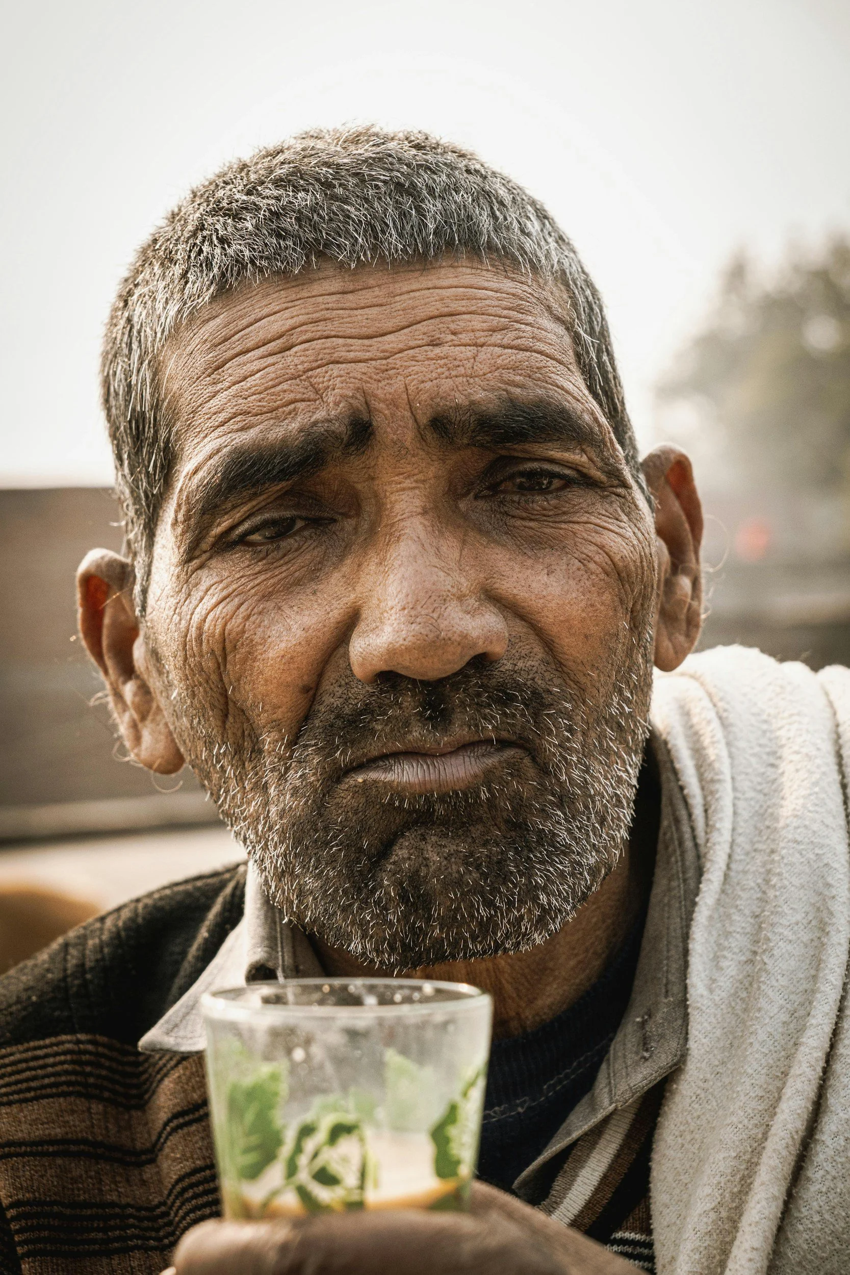 Close-up of an elderly man with grey hair and beard, holding a glass of tea, with a neutral background.