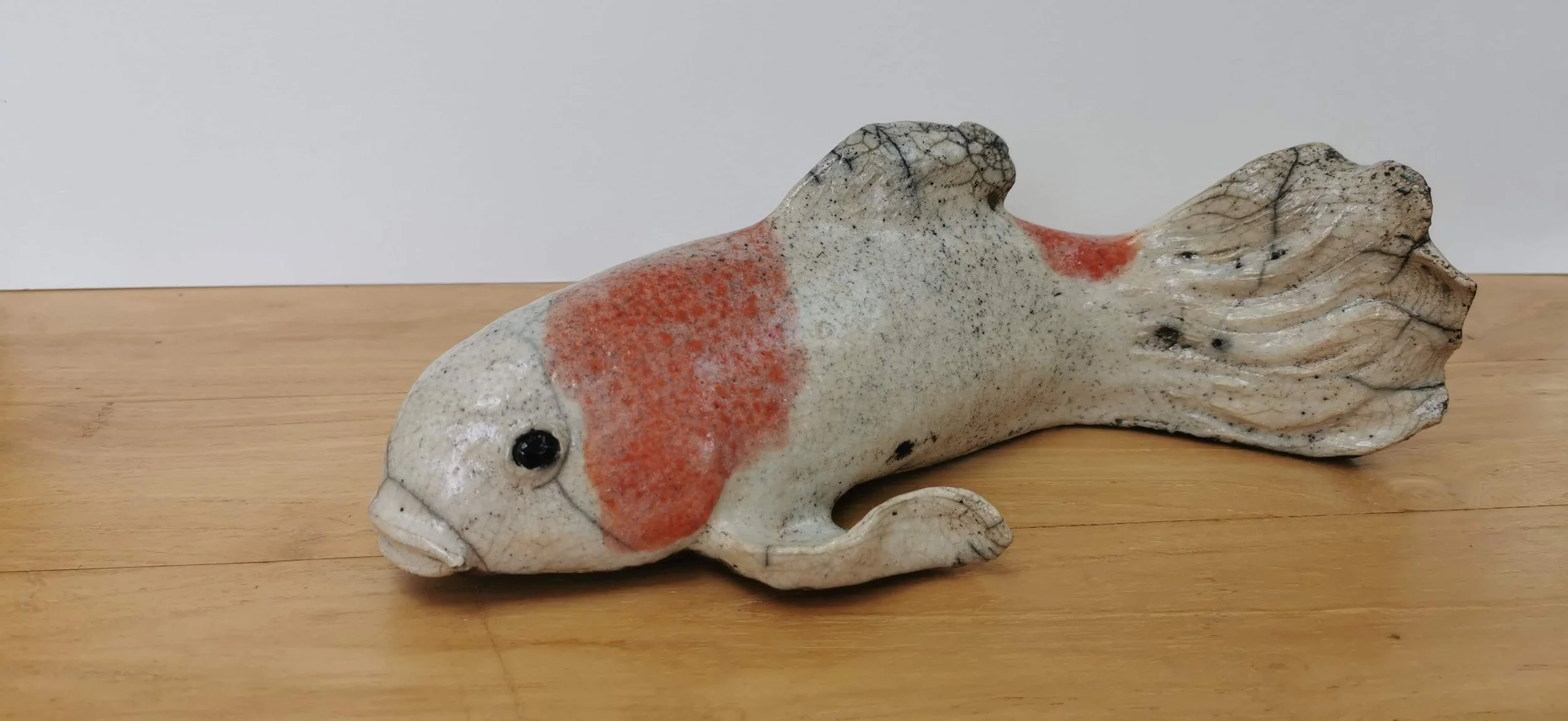 Statue en céramique d'un poisson rouge avec des nageoires et une queue détaillées, posé sur une surface en bois.
