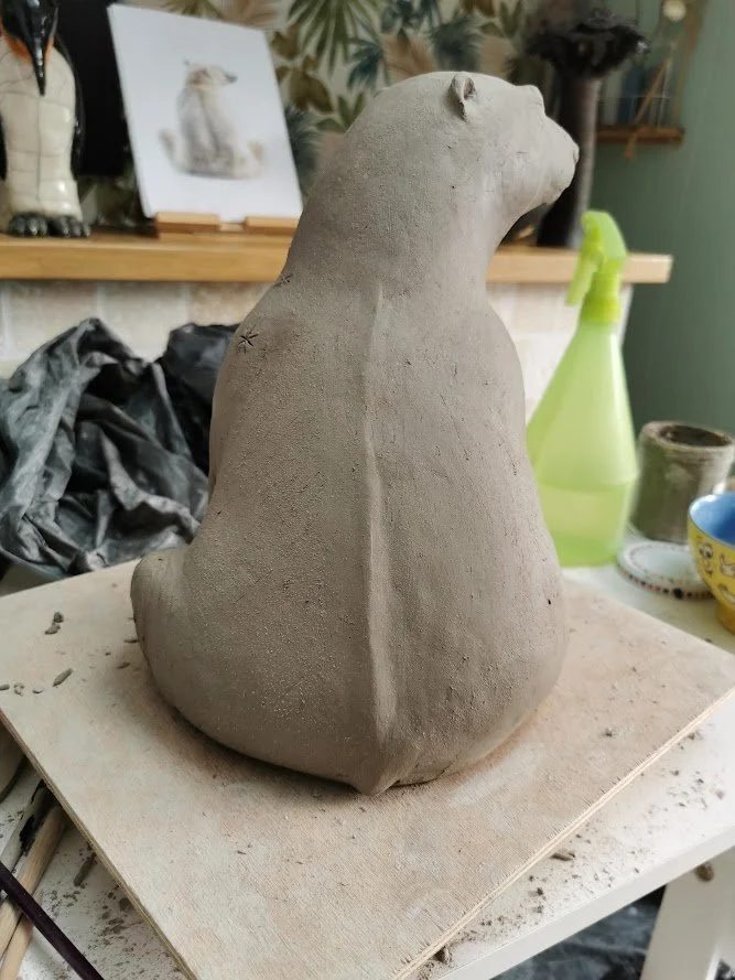 Sculpture en argile d'un ours, en cours de fabrication, placé sur une table de travail avec divers outils et objets en arrière-plan.