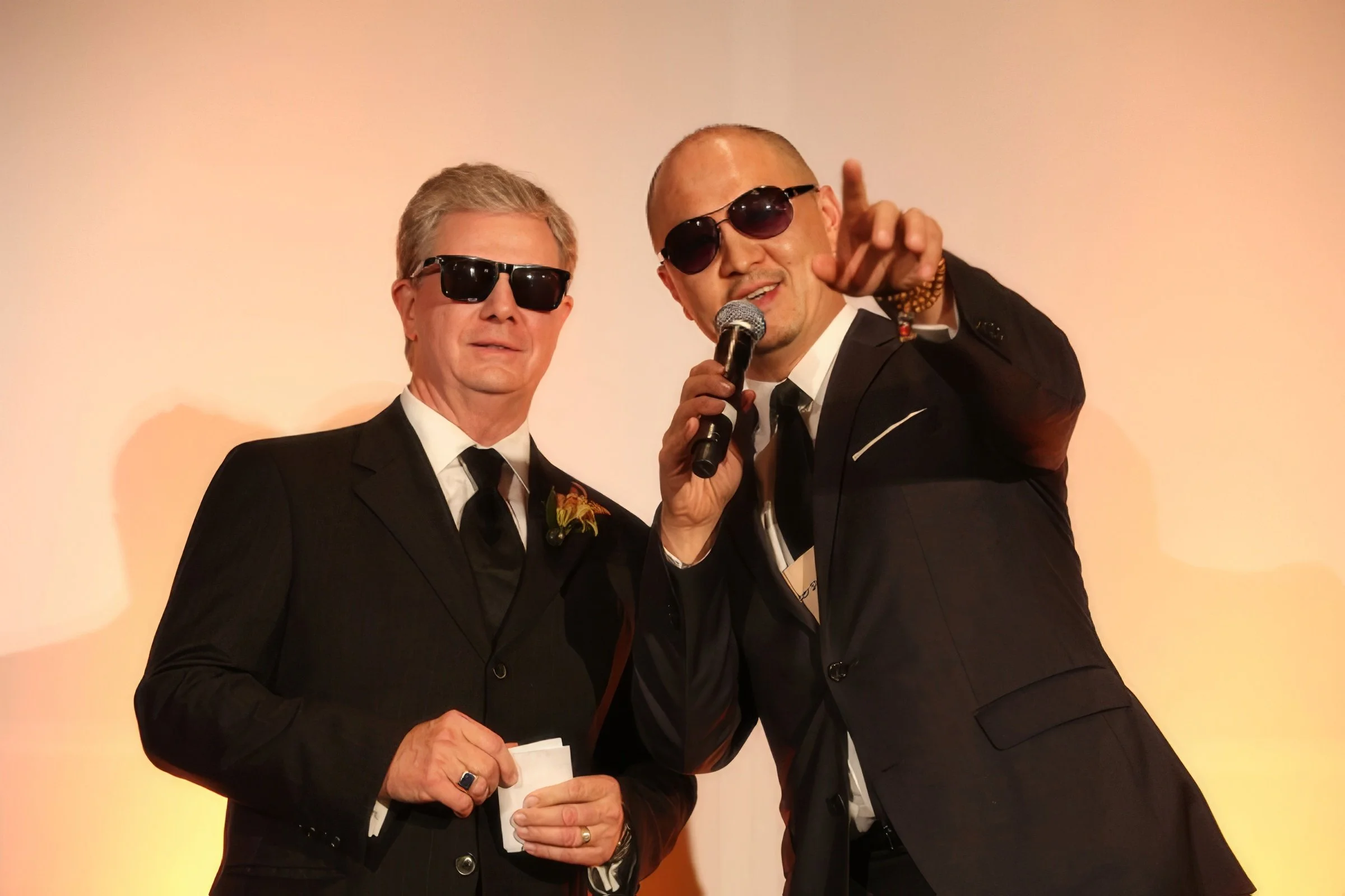 Two men in black suits and sunglasses, one holding a microphone, stand on a stage with a light-colored background.