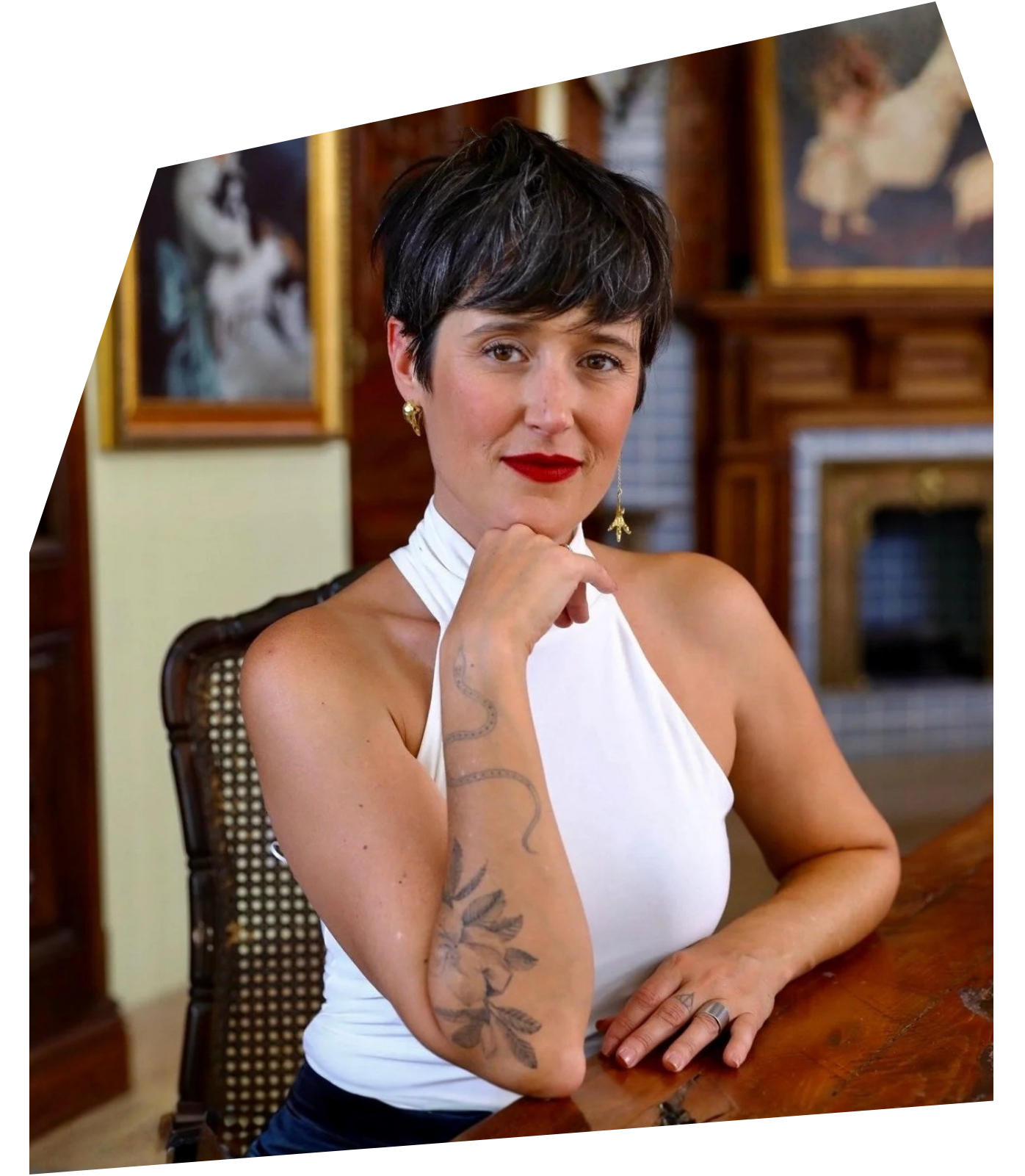 A woman with short dark hair and red lipstick sitting at a wooden table in an indoor setting with artwork and a fireplace in the background. She is wearing a white sleeveless top, gold earrings, and has a tattoo of a snake and floral design on her forearm.