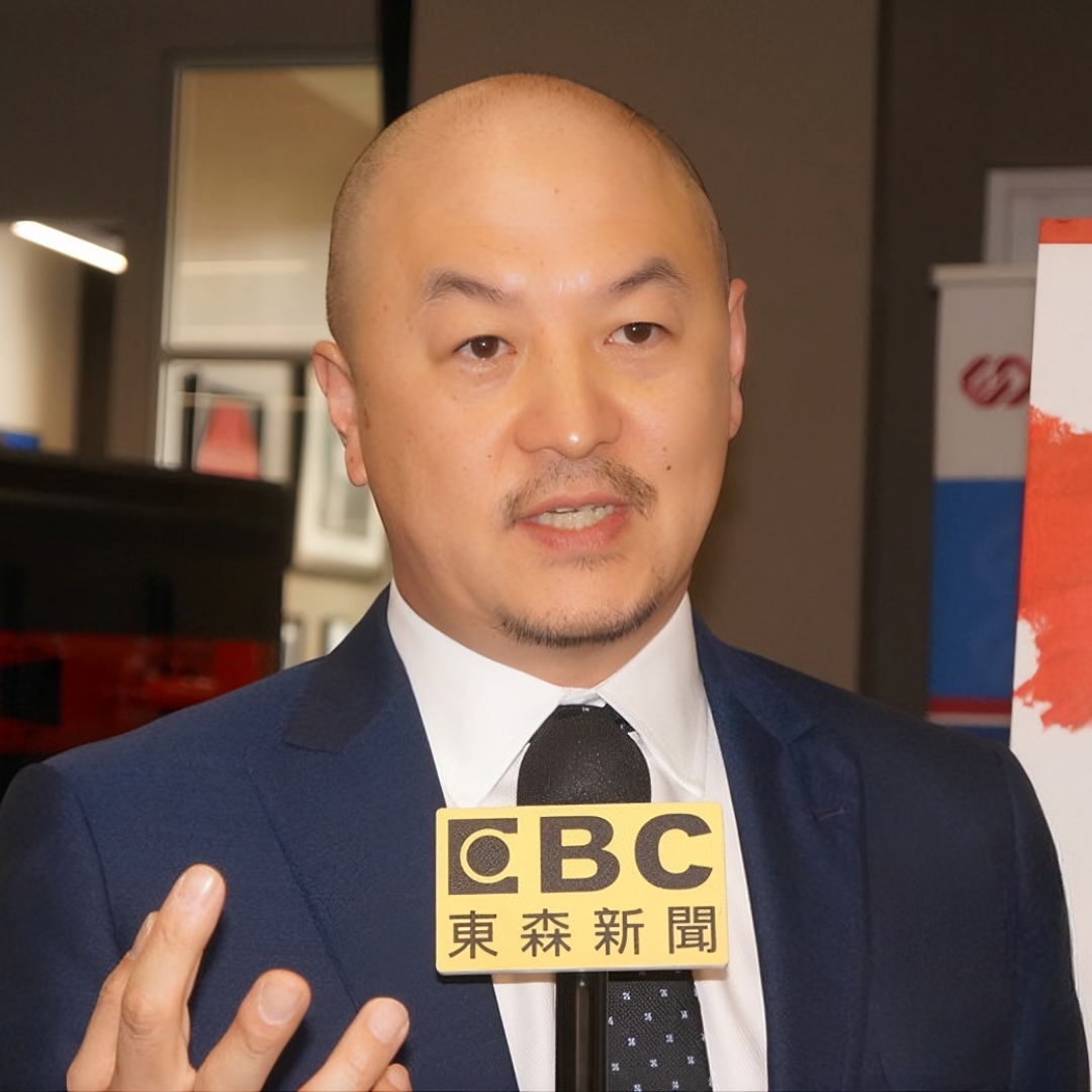 Man in a suit speaking into a microphone with a CBC logo at an indoor event.