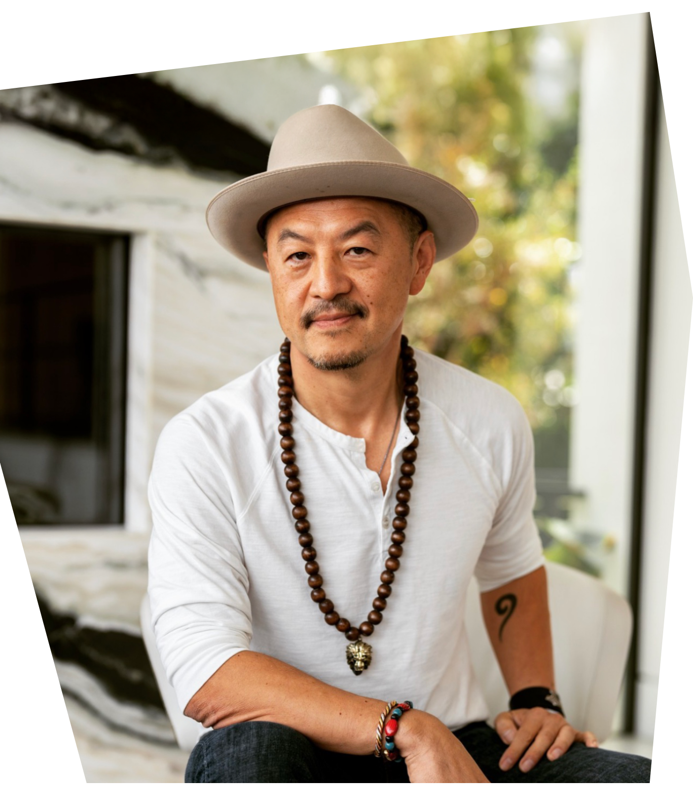 A man wearing a beige wide-brimmed hat, white shirt, and beaded necklaces sits indoors near a window with trees visible outside.