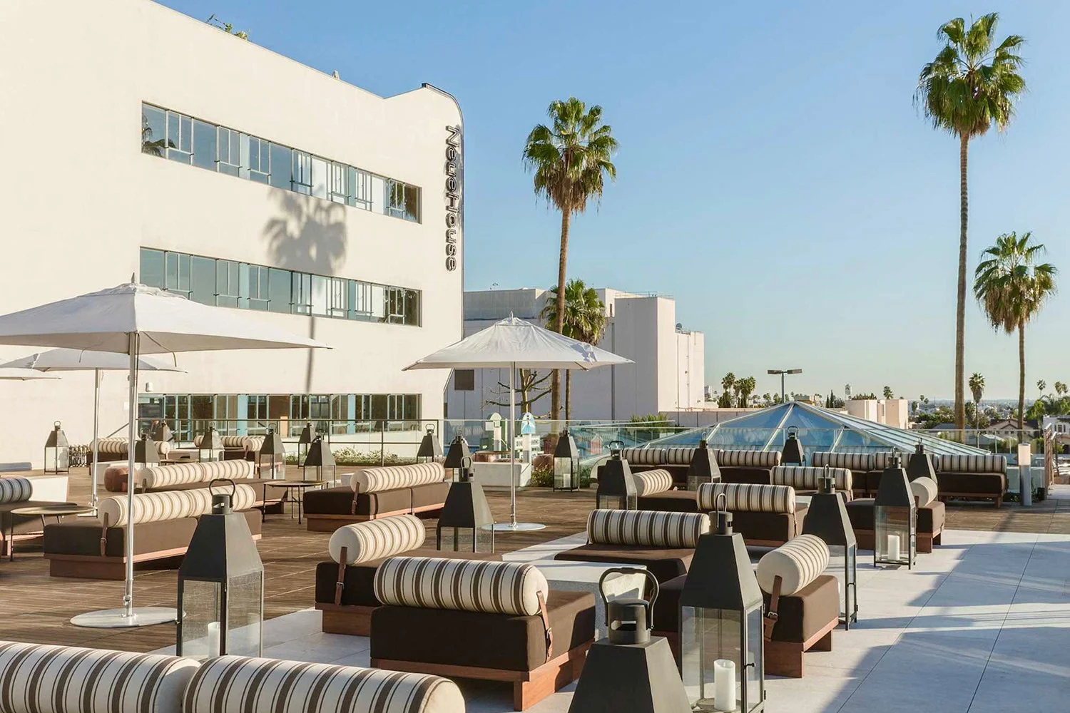 Rooftop lounge area with striped cushioned seating, white umbrellas, and lanterns, with palm trees and a white modern building labeled 'Neuman' in the background, under a clear blue sky.