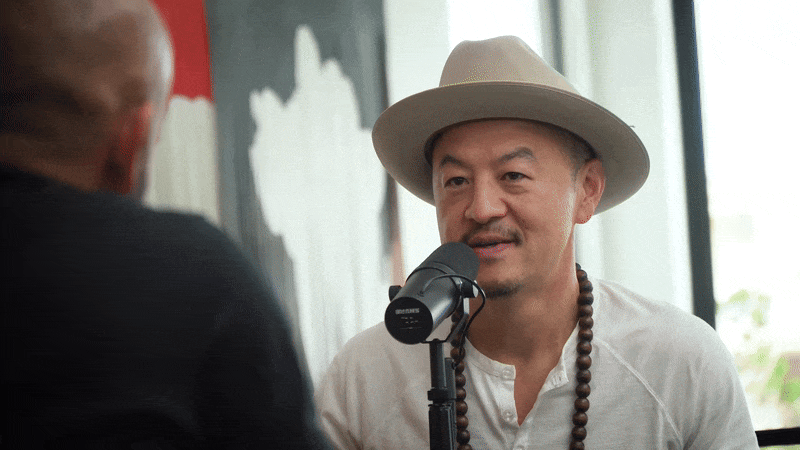 A man wearing a beige hat, white shirt, and beaded necklace speaks into a microphone during a podcast or interview, with another person partially visible in the foreground.