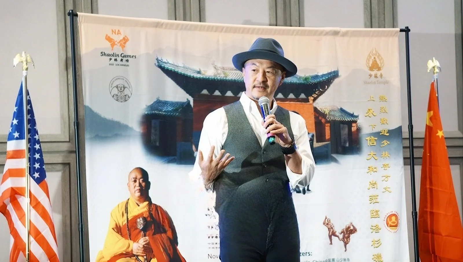 A man wearing a hat, white shirt, and vest speaking into a microphone on stage with flags and a banner behind him, featuring the American and Chinese flags, and images of a monk and traditional Chinese architecture.
