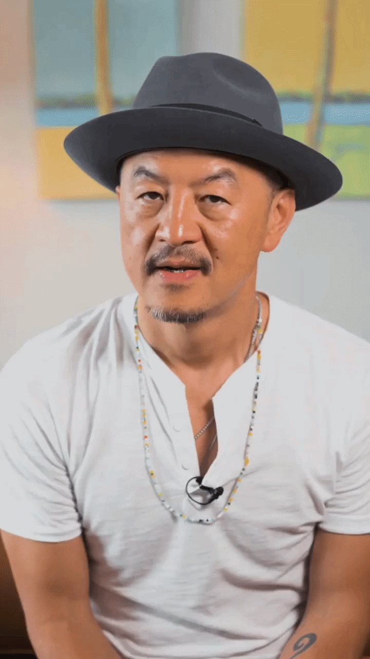 A man wearing a dark gray hat, white shirt, and beaded necklace, sitting and talking to the camera indoors with a colorful abstract painting in the background.
