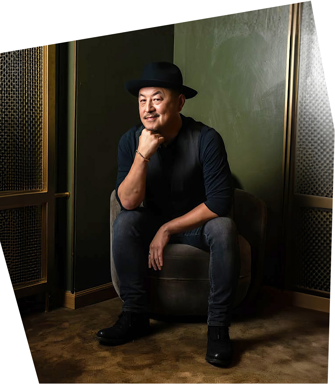 A man wearing a black hat, black shirt, and dark pants, sitting on a grey chair in a dimly lit room with green walls, posing with his chin resting on his hand and smiling slightly.