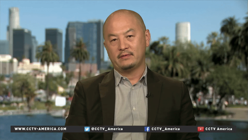 Male news anchor with short hair and beard, wearing a dark blazer and light gray shirt, speaking in a news interview with a cityscape background including tall buildings, palm trees, and a body of water.